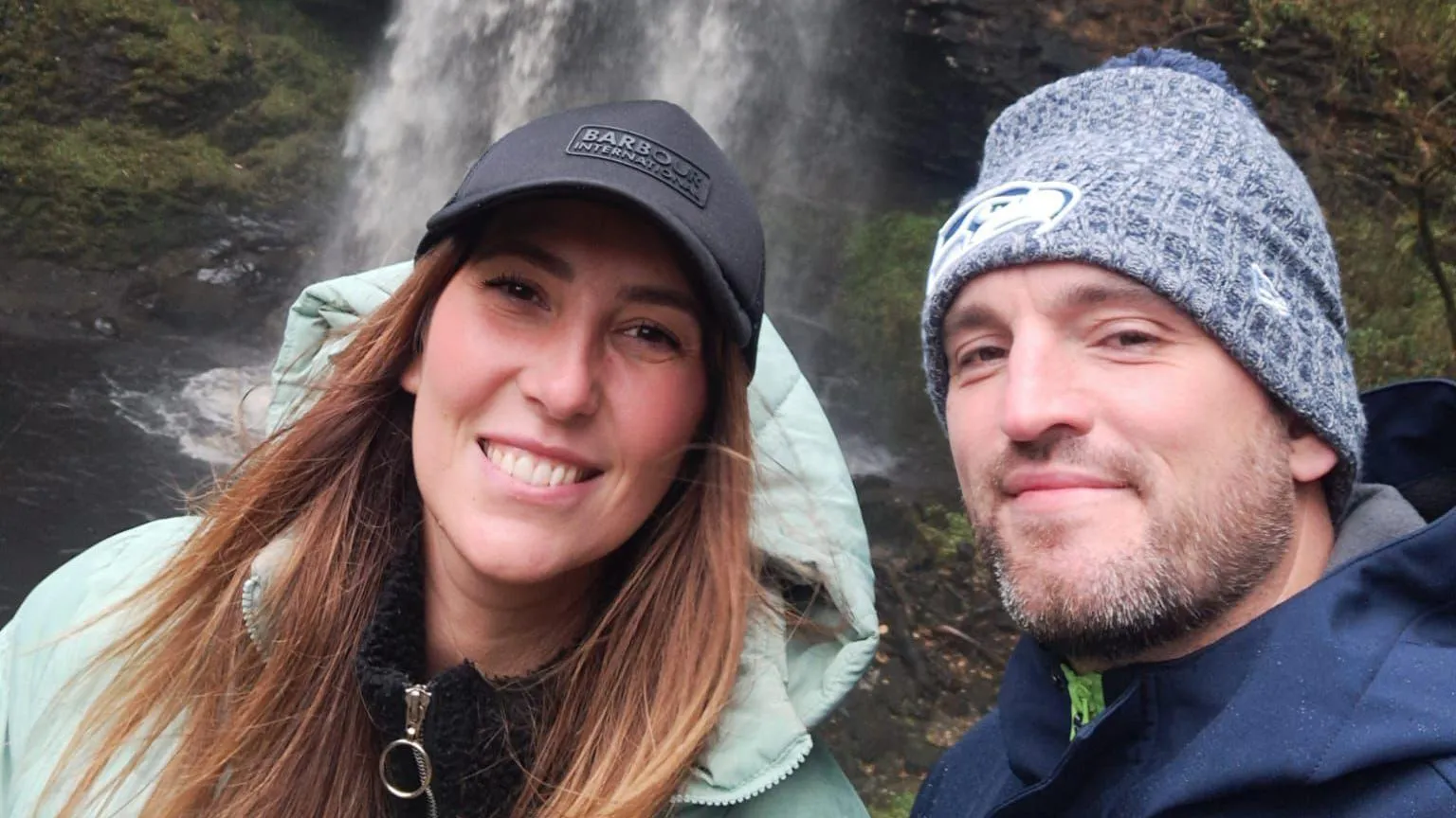 Leah Matthews A selfie of the couple - the woman is wearing a black cap with long dark hair, with a light blue raincoat, the man a grey beanie. He has short greying stubble and is wearing a blue waterproof. They are smiling and in the background is a waterfall.