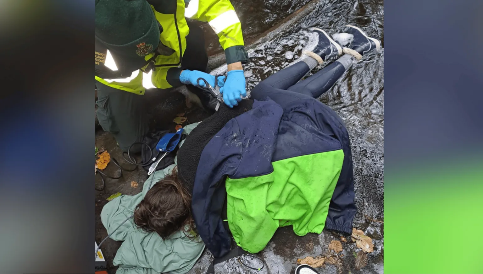 Leah Matthews A person is lying on the floor with a coat over her. Her feet are in water. A paramedic is close to her and is cutting away some of her clothing with scissors.
