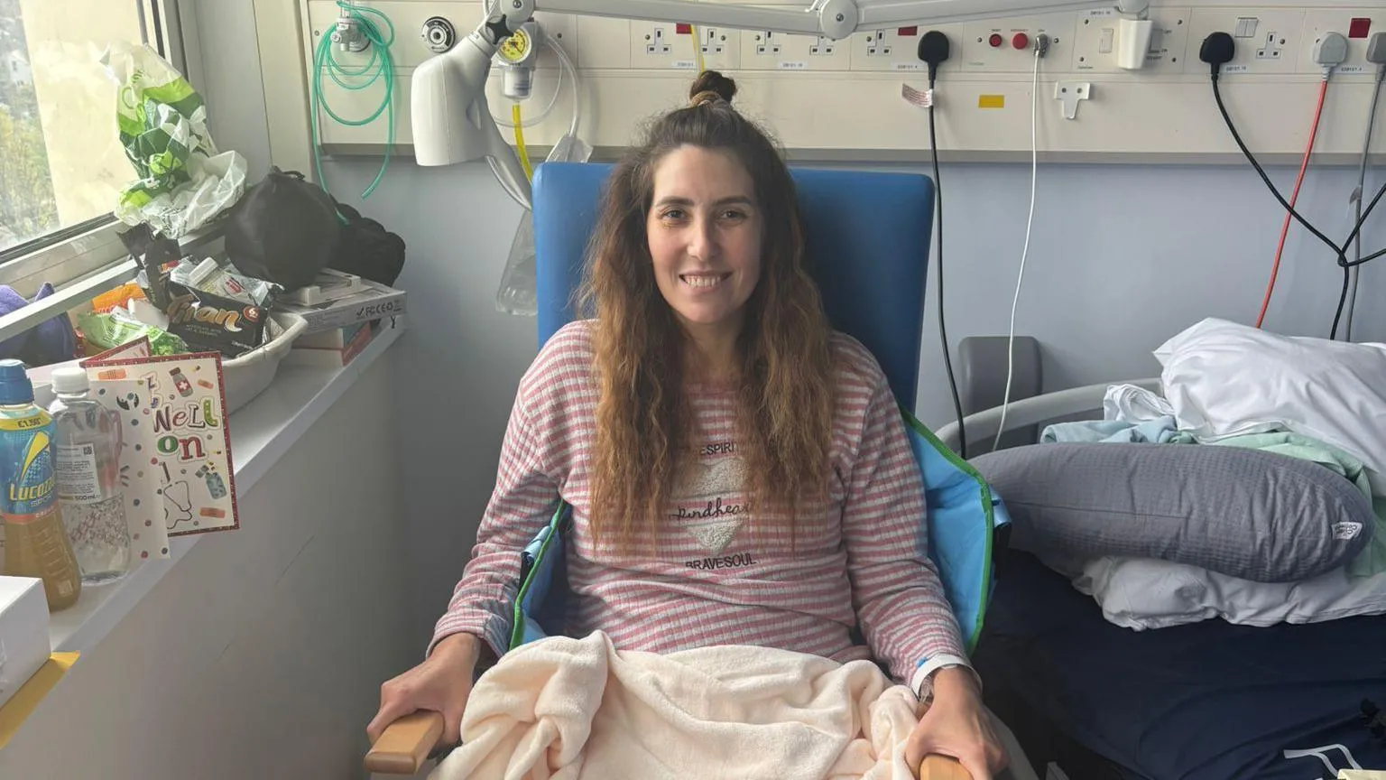 Leah Matthews Leah sat in a chair next to a hospital bed. She has cards and drinks next to her and is smiling. She has a blanket over her legs and is wearing a pink stripey top. Her long wavy hair is half tied up. 