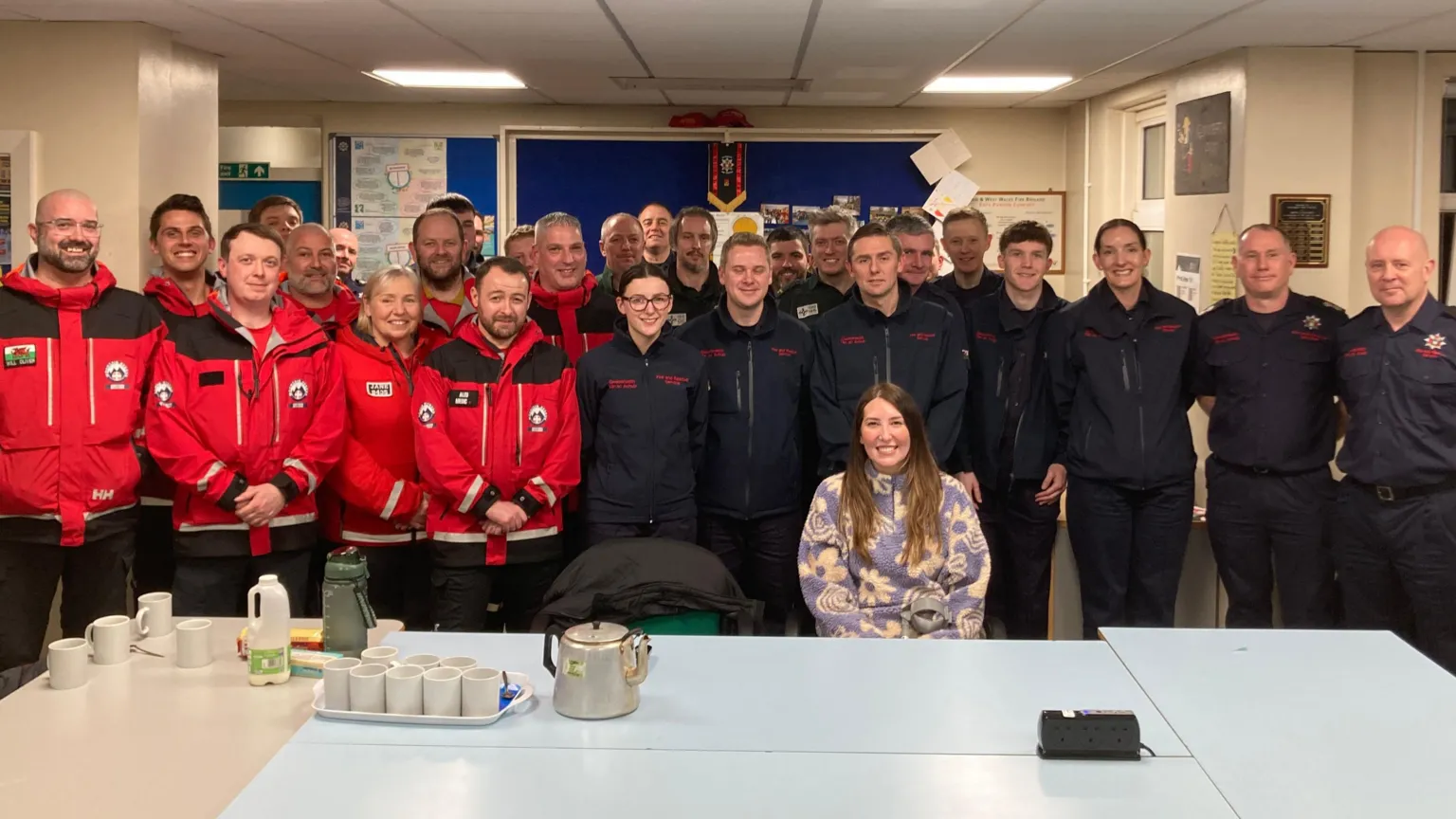 A large group of people - members of Western Beacons Mountain Rescue Team, Mid and West Wales Fire Service and the Welsh Ambulance service with Leah Matthews. She sits down by a table with the three groups behind her, one group in red jackets, and the others in navy blue. On the table is a teapot, milk carton and mugs. 