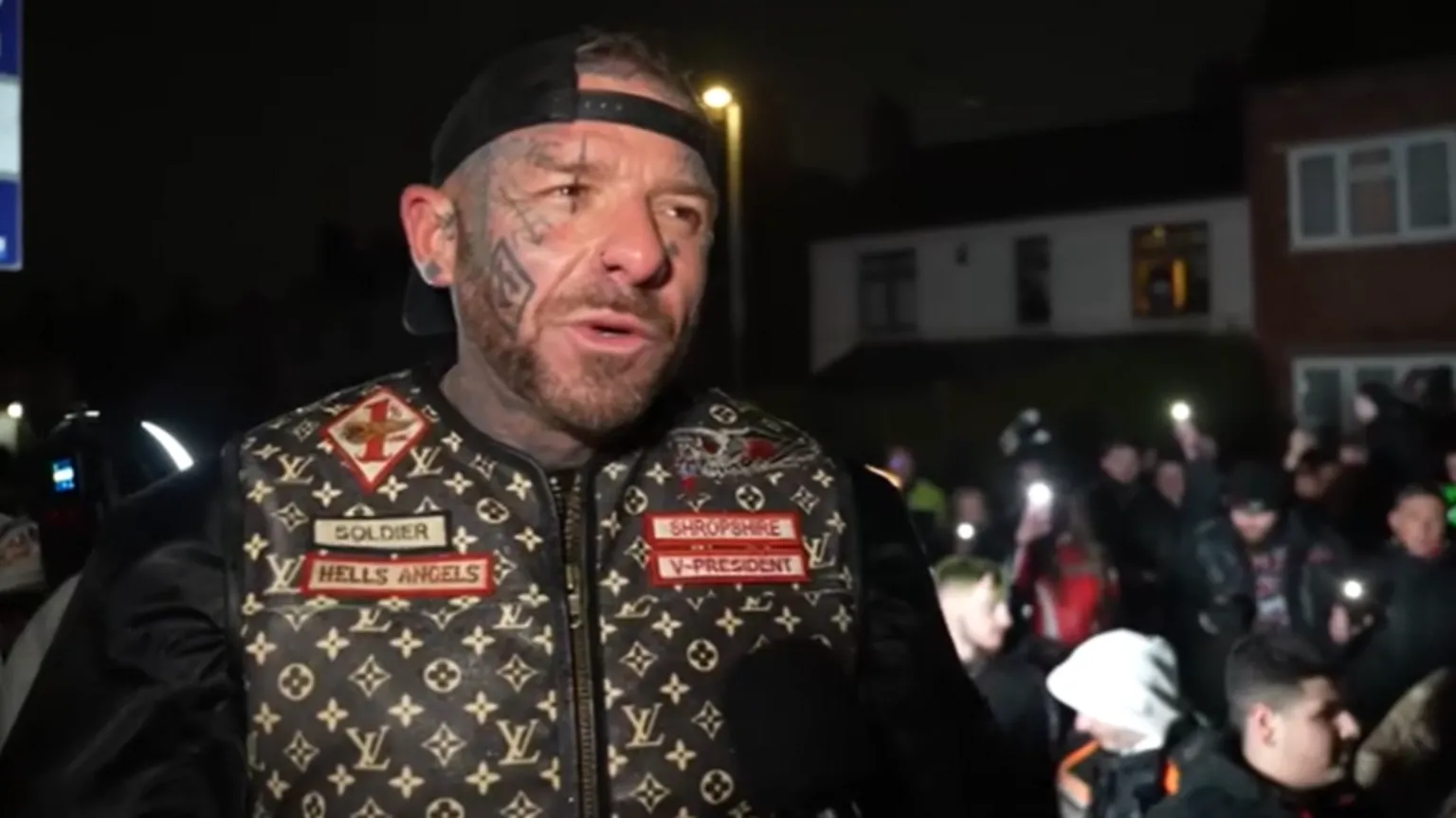 A man wearing a biker jacket with and backwards cap talks to a camera in a residential street. He has tattoos on his face.