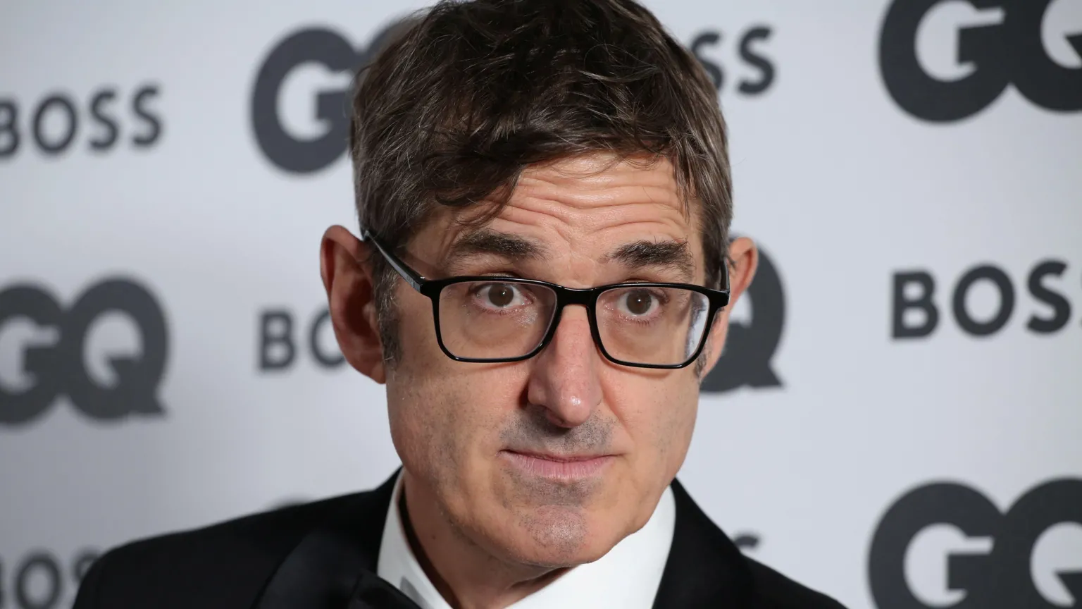 Lia Toby/ Louis Theroux is wearing a tuxedo. A backdrop is behind him. He is wearing black glasses. 
