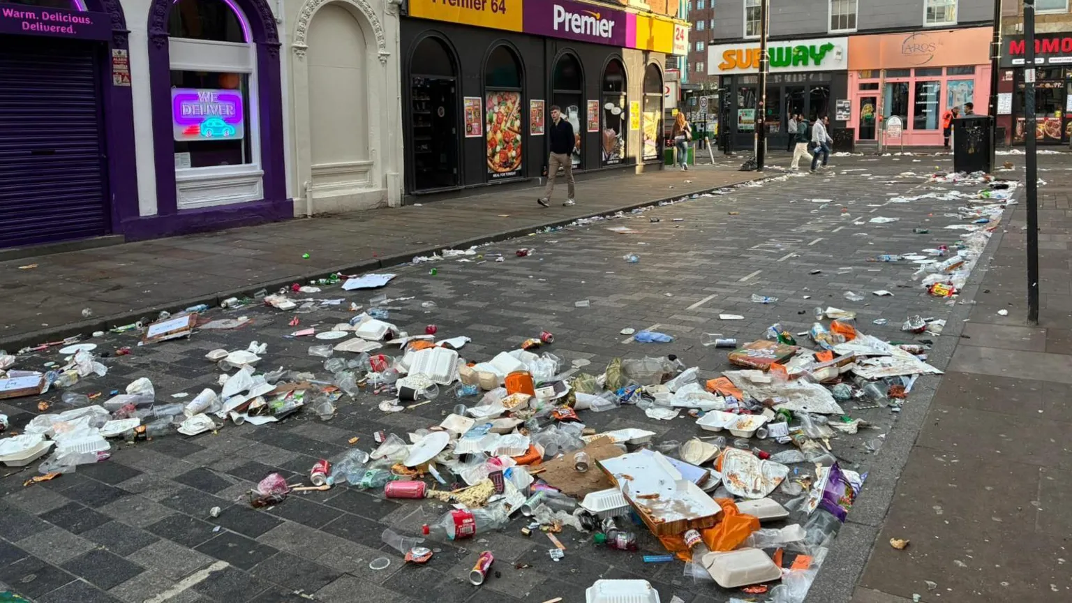 Litter is sprawled across a paved road with pedestrians walking by. The street also has shops on it.