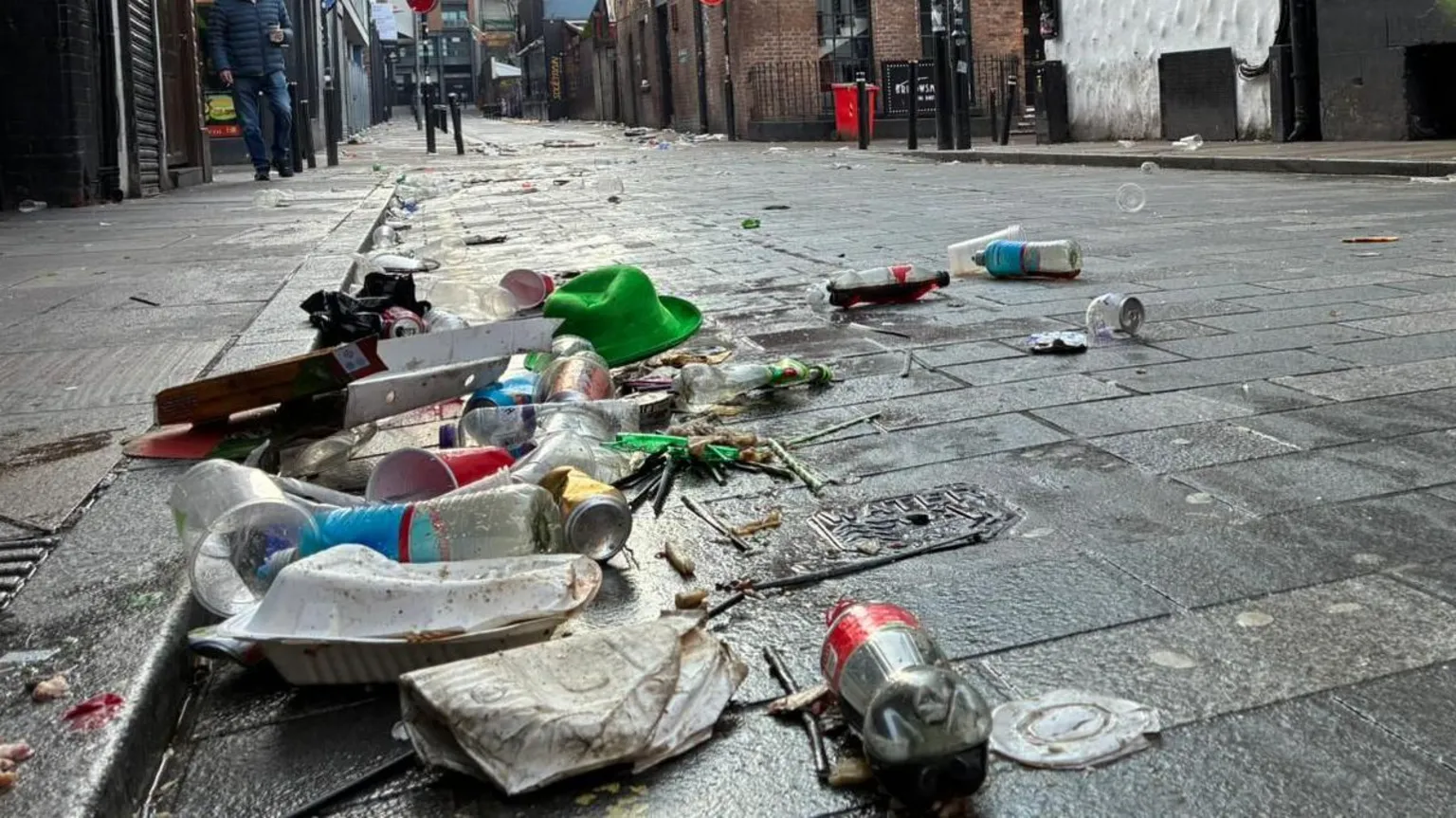 A close up of a road with a pile of litter next to the curb. The litter includes plastic bottles and cups, food boxes and a green bowlers hat