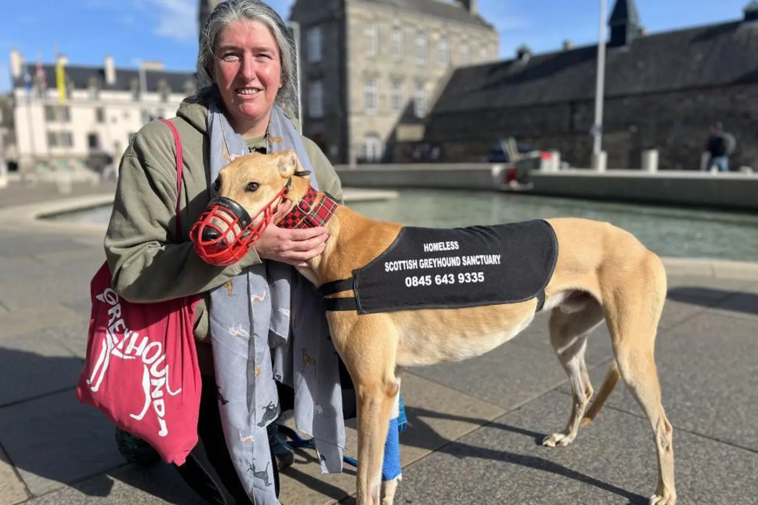 A woman with grey hair kneels beside a greyhound, which has a black rug on with Scottish greyhound sanctuary branding. There is a pool of water and stone buildings in the background. 