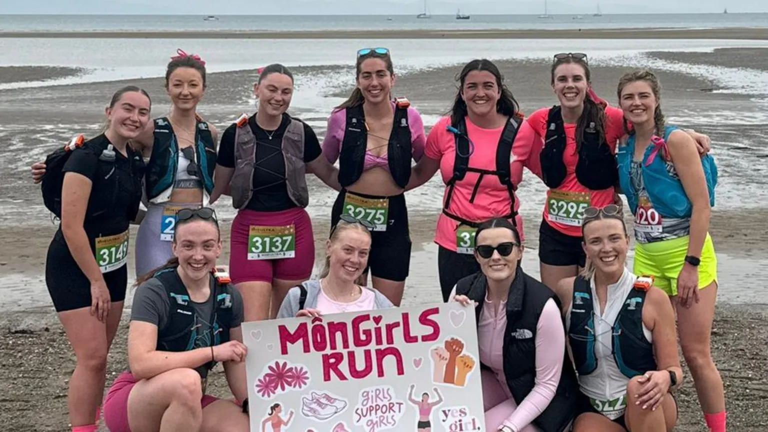 Gwen Owen A group of female runners posing in a group photo on a beach with a Môn Girls Run poster