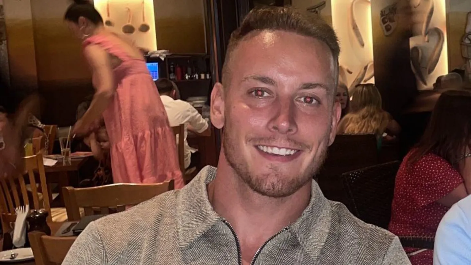  George, a man with short light brown hair and wearing a grey collared top with herringbone design fabric is smiling. He is sitting at a table in a restaurant.