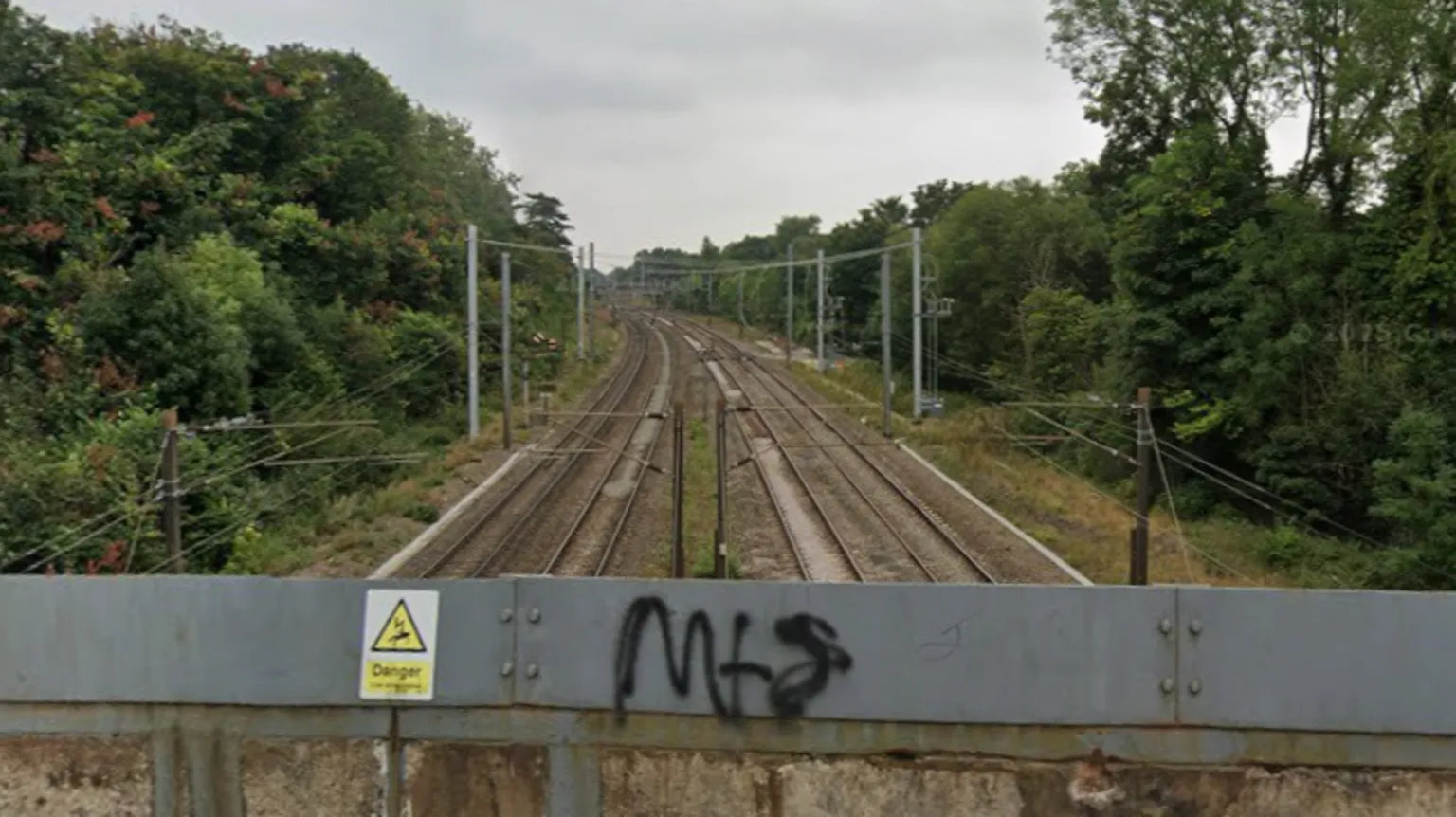 Google A railway line with four tracks and overhead cables. There are trees either side of the line. In the foreground there is a grey railing with a danger sign indicating electric cables.