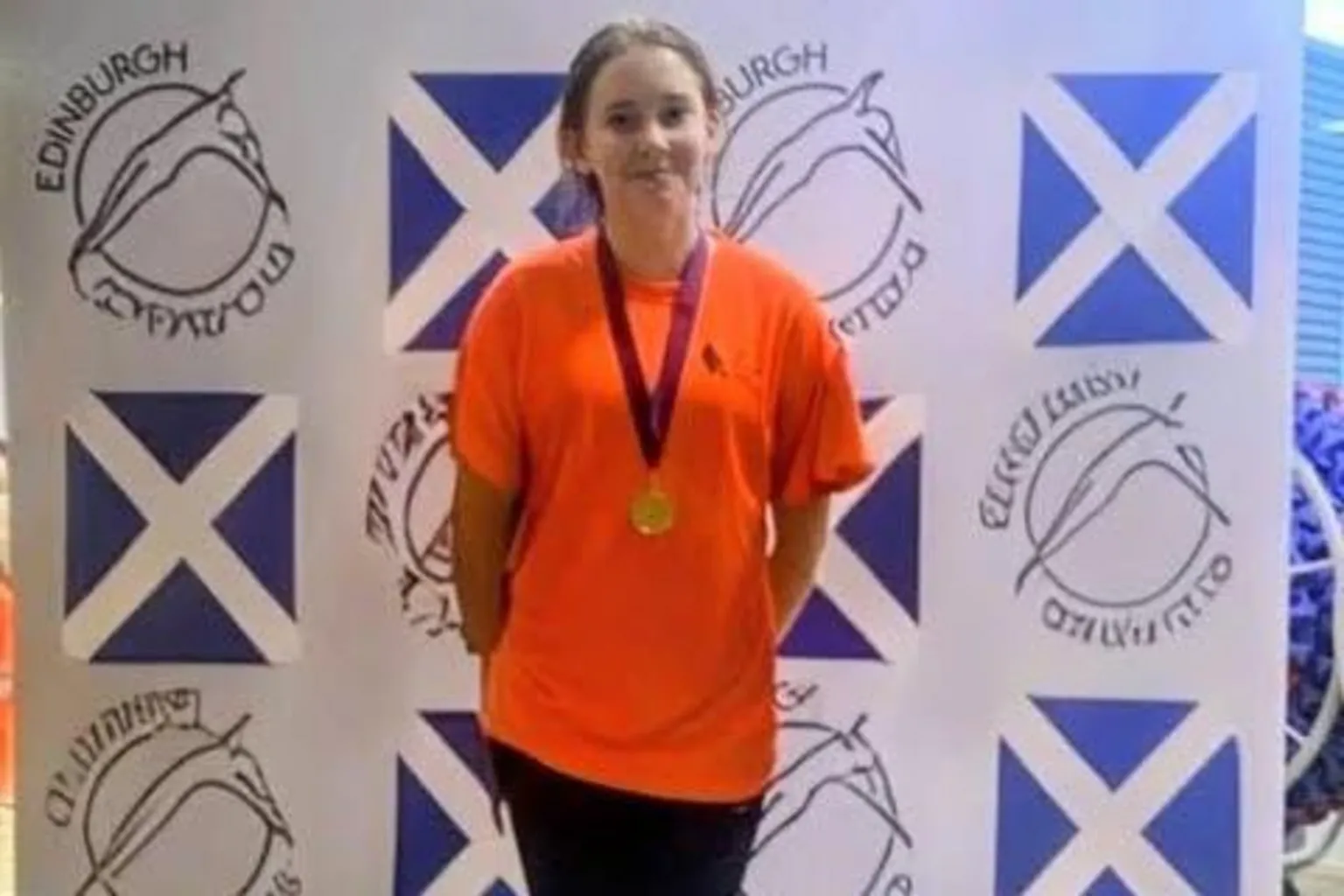 Heather Graham A young teenage girl wearing an orange t-shirt with a gold medal around her neck stands on a podium.