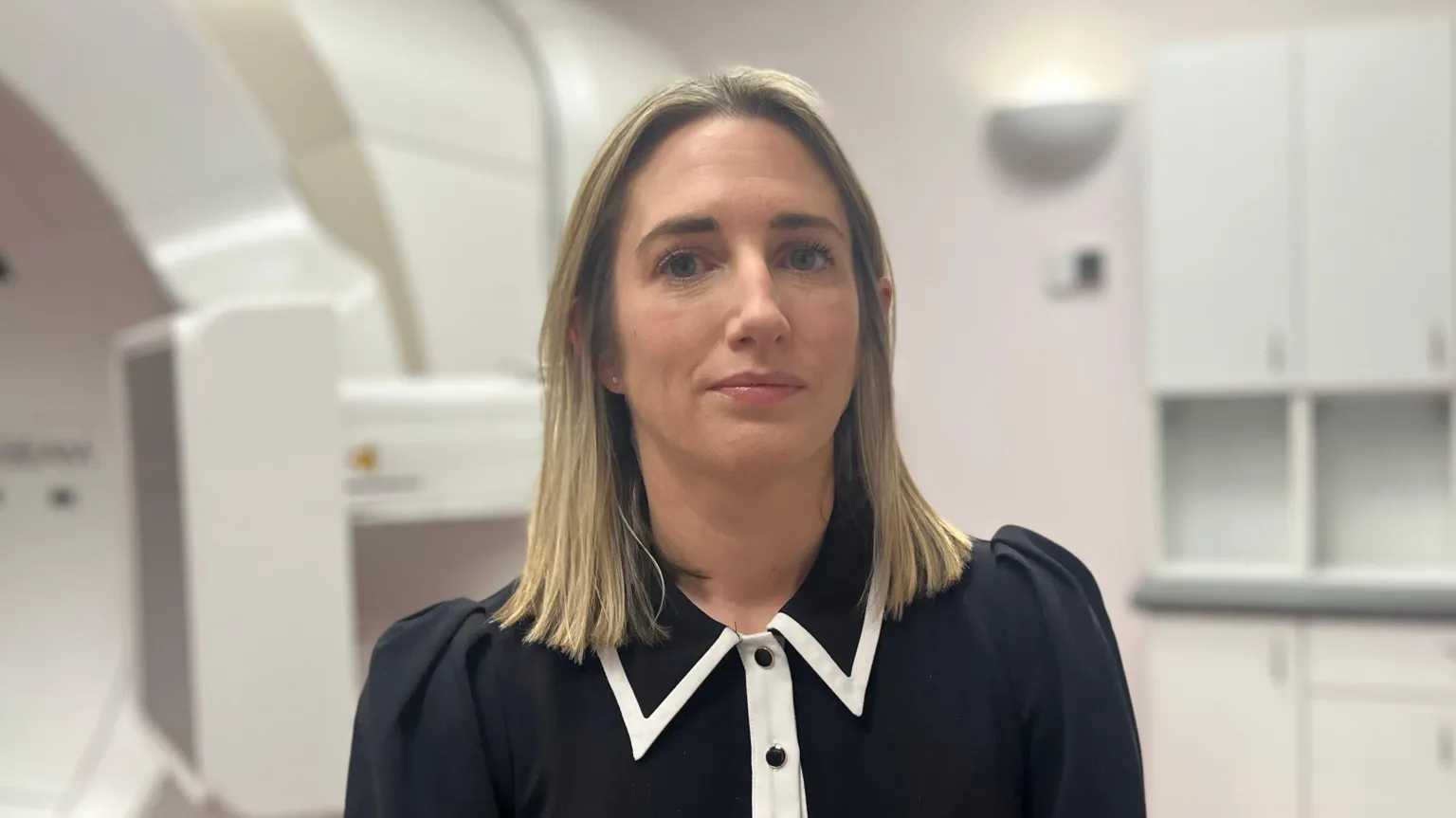 Dr Hanna has shoulder length blonde hair and is wearing a black top with white trim and standing in a hospital room, behind her the edge of a large white medical scanner can be seen.