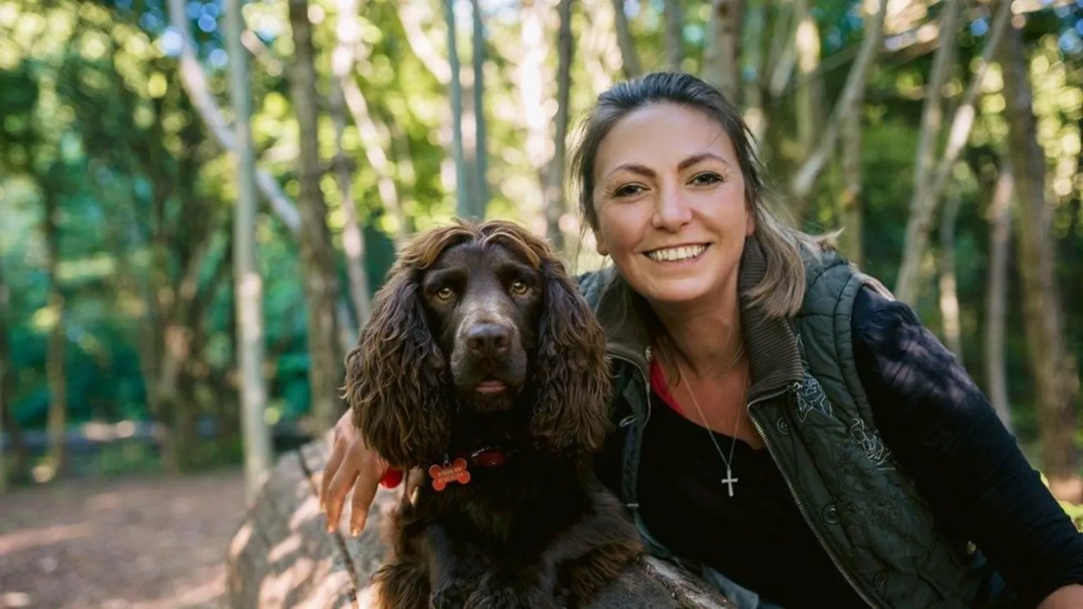 Hannah Molloy Hannah Molloy, a woman with her tied back, wears a dark top and dark green quilted gilet. She is smiling at the camera and has one arm resting on brown spaniel Falkor, who wears a red collar and is laid on a log in a forest.