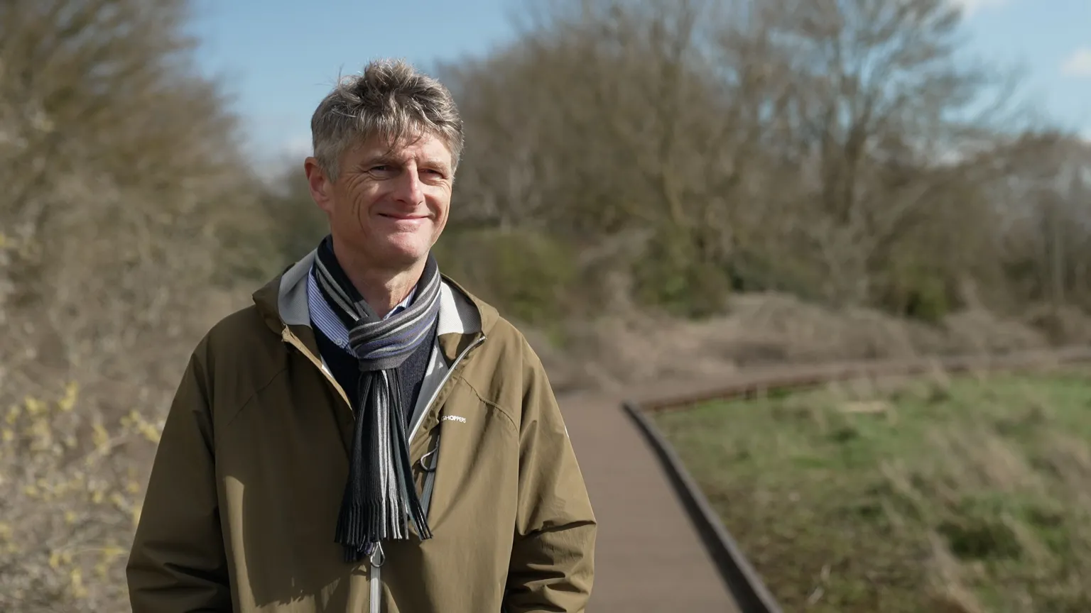 BBC / Kevin Church A man with silver hair and a stripey scarf weaing a khaki jacket is standing with a smile on a wooden boardwalk. Trees in the background have no leaves and the piece of land next to the walkway has some green grass but is mostly mud. 