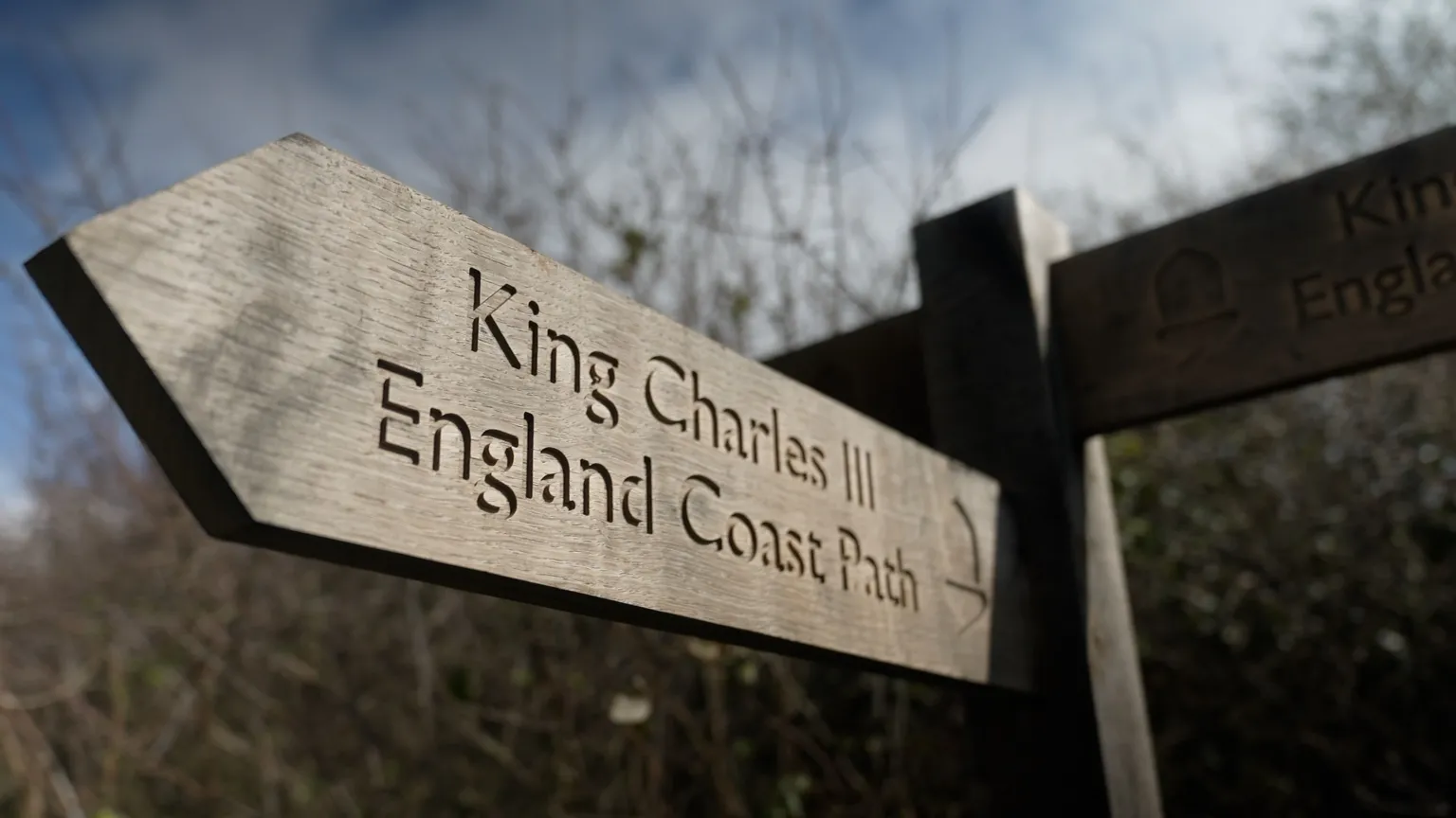 BBC /Kevin Church A wooden sign shows letters - King Charles III England Coast Path with some trees without leaves in the background against a blue sky with some clouds. 