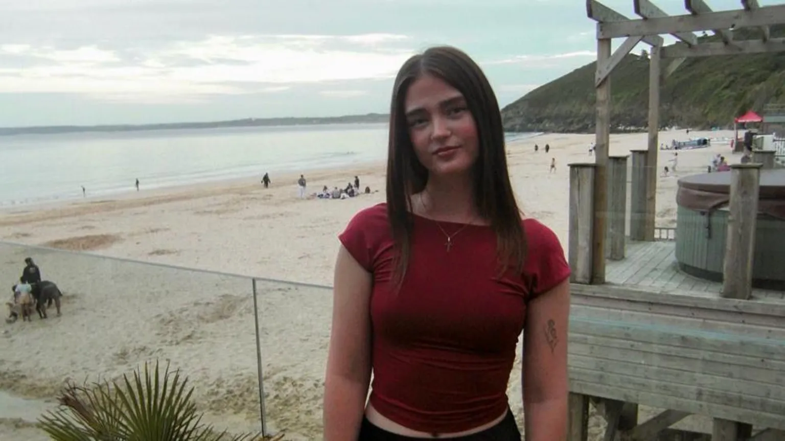 Family Tyra Skinner is standing in front of a sandy beach with a beach hut behind her. She is wearing a red T-shirt and a necklace and has long, brown hair.