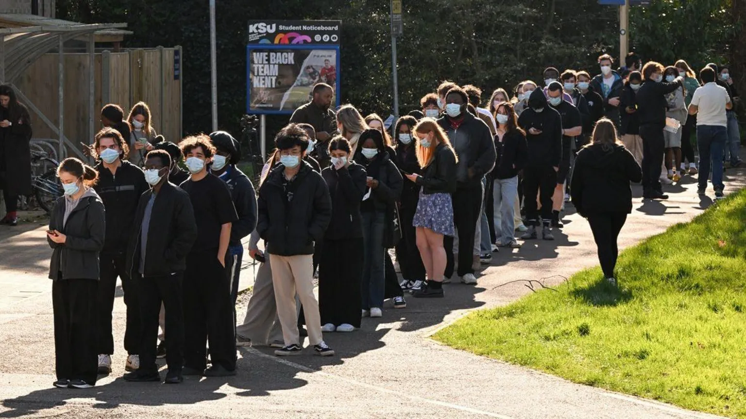  People queue to receive vaccinations at the sports centre at the University of Kent campus in Canterbury. They are all wearing masks and are lined up in the sunshine near an area of grass. There are trees behind them and signposts and there is a student noticeboard with a poster.