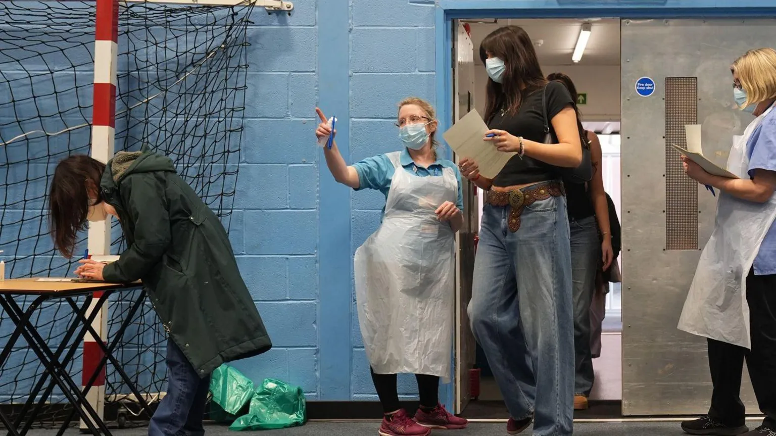  Students are walking into a sports hall carrying paperwork. Staff are near the door wearing white aprons, dealing with forms and telling people where to go. A girl in jeans and trainers and a coat is completing a form on a table near a sports hall net. The walls are blue.