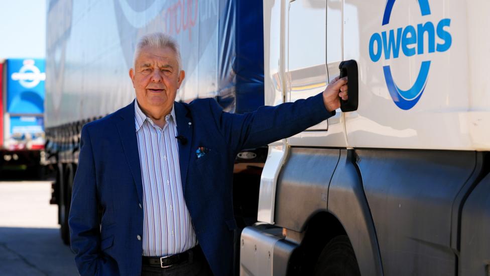 Dyn mewn siaced glas tywyll a chrys golau yn sefyll wrth ymyl lori gywn a glas o gwmni Owens