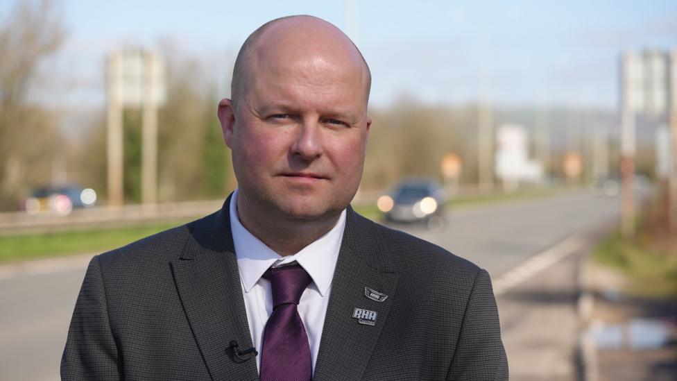 Dyn moel mewn siwt tywyll, a tei porffor, yn sefyll wrth ymyl ffordd ar ddiwrnod heulog.