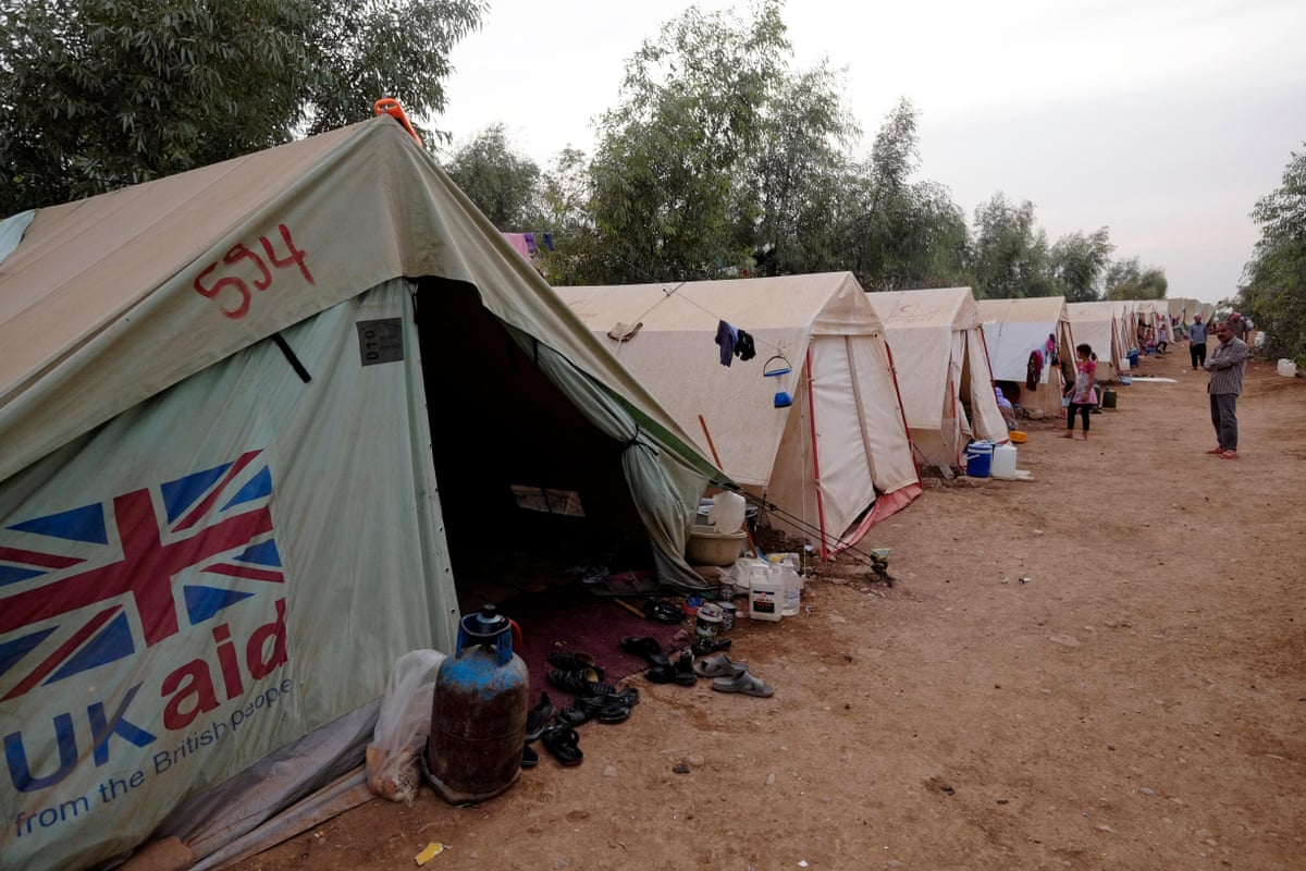  A line of white tents pitched on a sandy surface: a union jack flag and the words UK aid from the British people are seen on the side of the tent closest to the camera. There are shoes, water containers and cooking gas cylinders at the tent's opening.