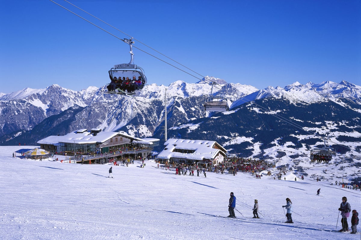 a chair lift passes over a ski slope leading down to restaurants with mountains behind
