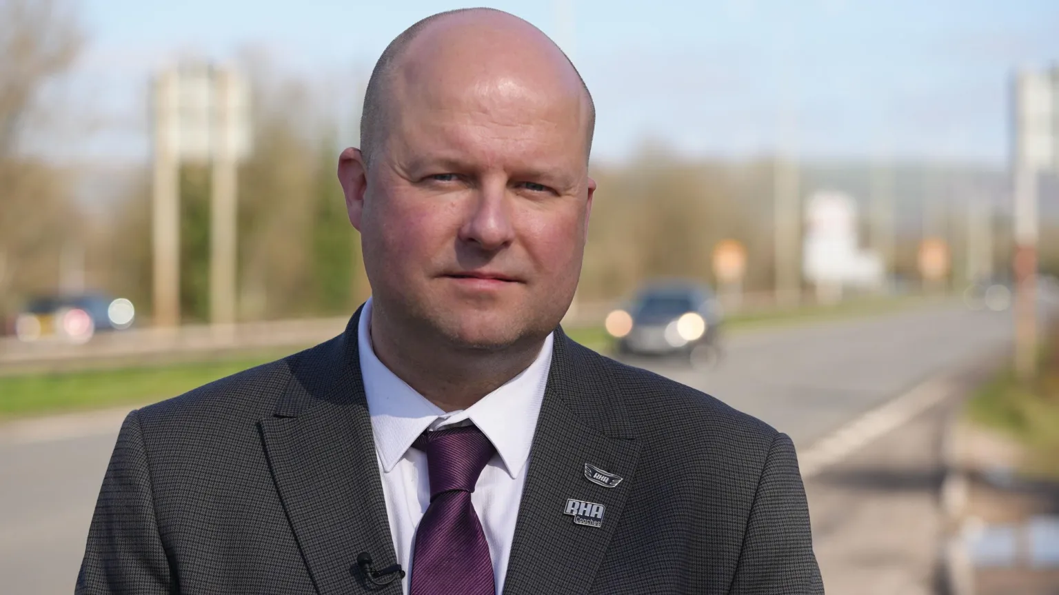 Geraint Evans, a bald man in a dark grey suit and purple tie, is looking into the camera. It's a sunny day, and he is standing at the side of a dual carriageway in Pencoed, which is blurred in the background. 