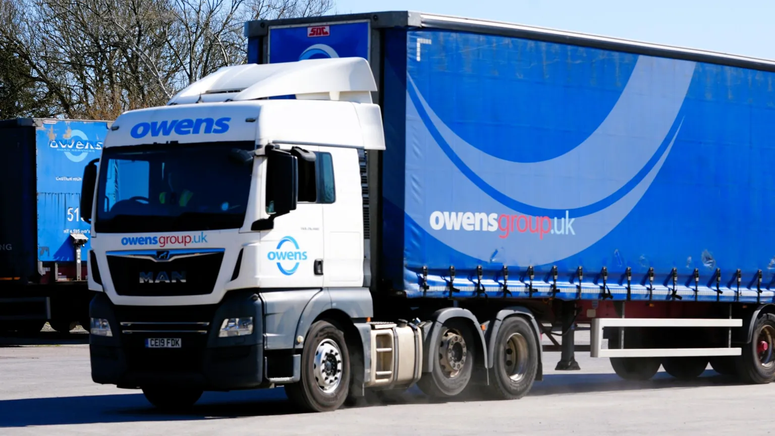 A blue Owens Group trailer is pulled by a white tractor unit on an industrial site, with other blue trailers parked in the background.