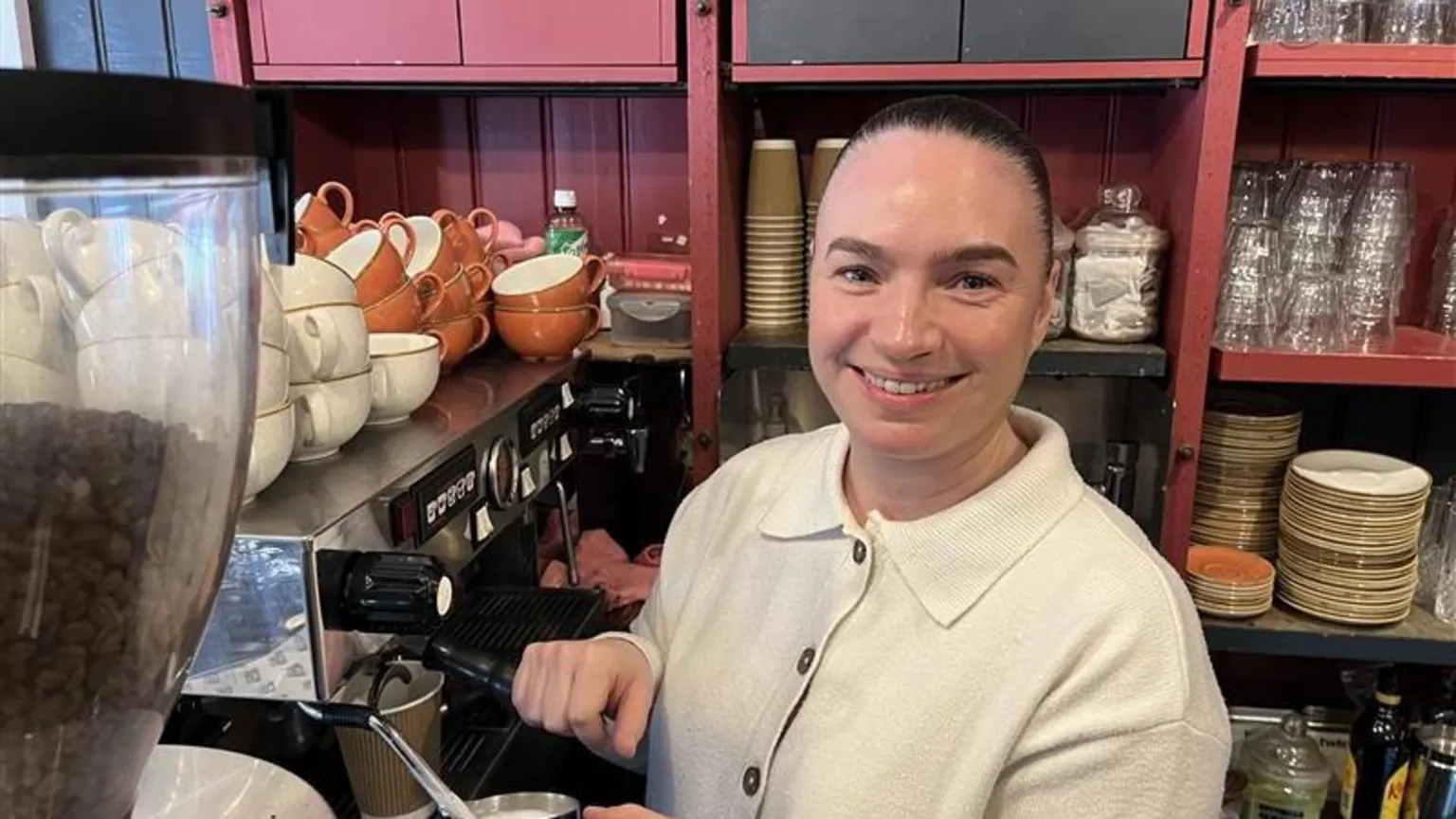 Phil Harrison / BBC A woman wearing a white top stood in a pink cafe next to a silver barista-style coffee machine.