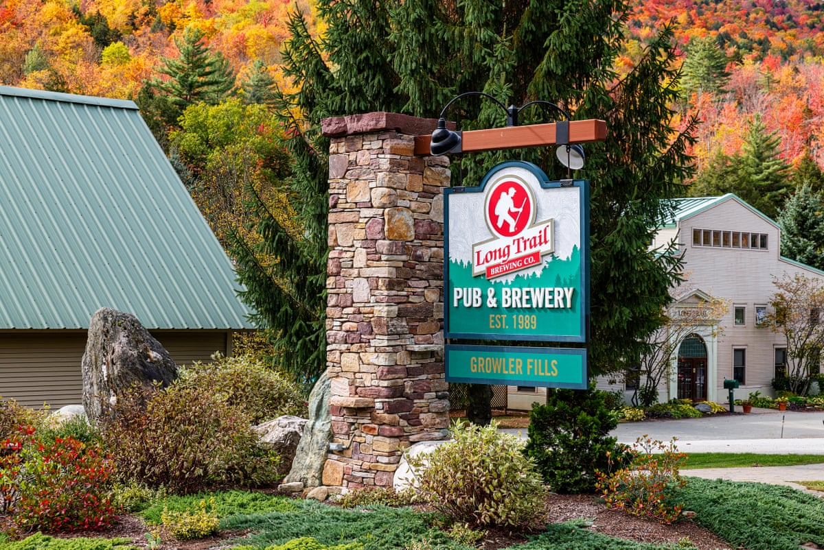 A sign – saying 'Long Trail Brewing Co Pub & Brewery Est 1989 Growler Fills – hangs outside a building surrounded by lush vegetation with fall colors