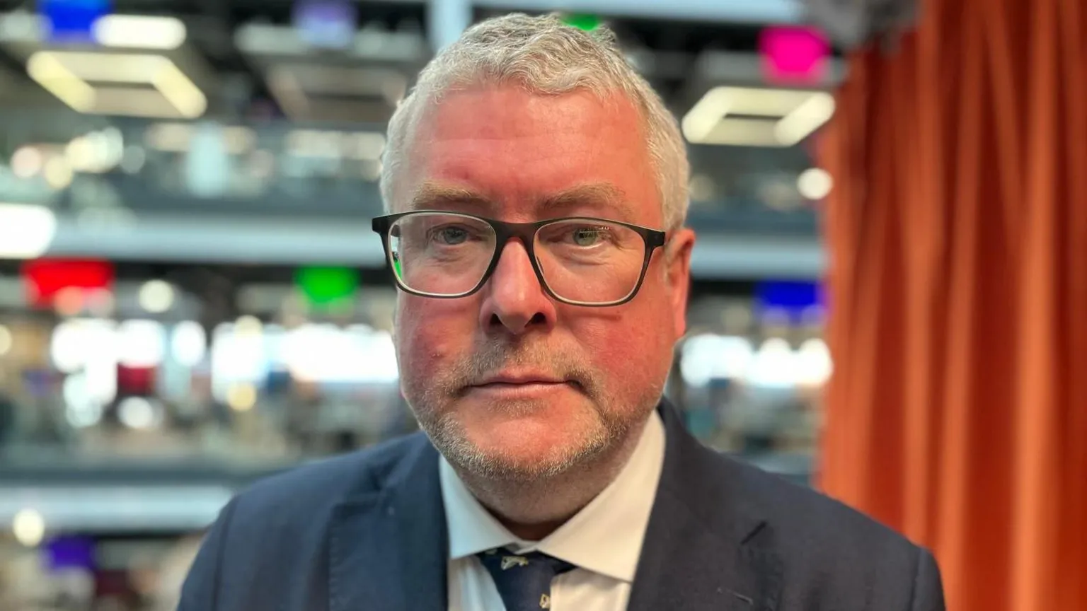 Prof Barry has white, short hair and is wearing dark-rimmed glasses. He has a navy suit, white shirt and navy tie on. He is stood with the BBC offices blurred in the background.
