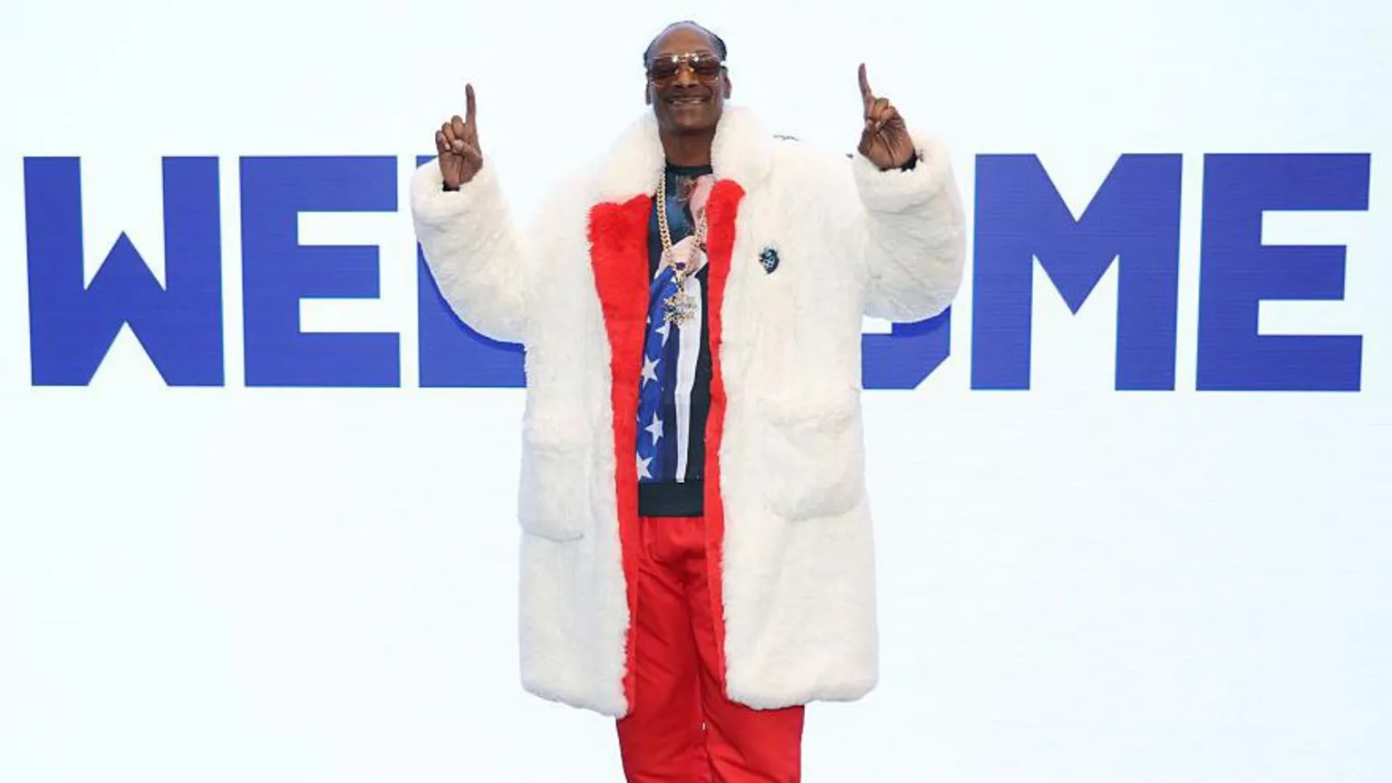 Snoop Dogg wears a fur coat over a red tracksuit, fingers pointed upwards while standing in front of a large sign saying 'Welcome' at the Winter Olympics
