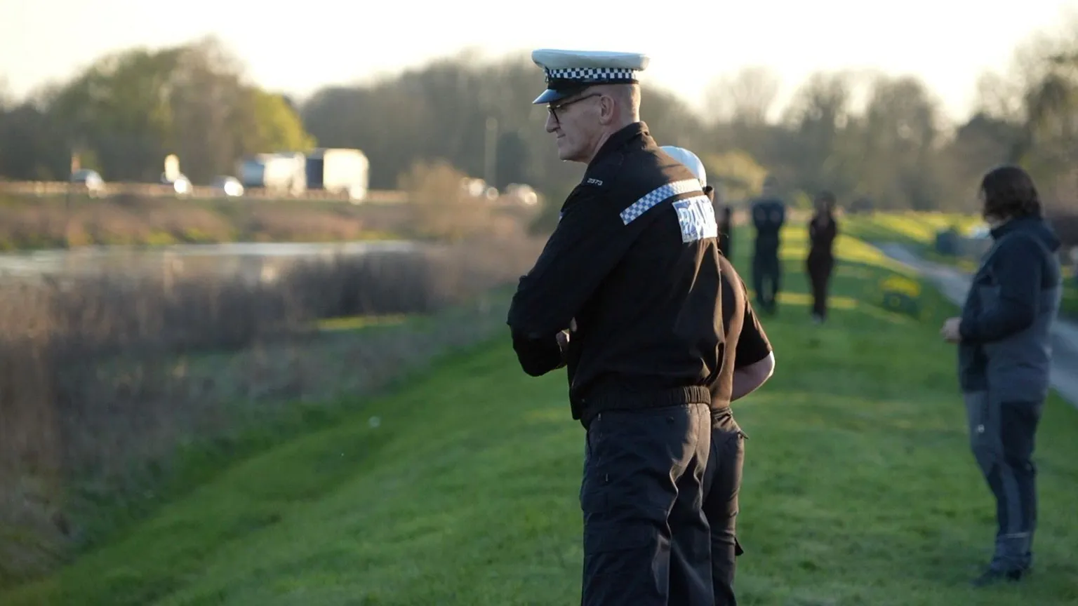 Qays Najm/BBC A side-on view of a police officer in black uniform, wearing a white and blue police flat cap, standing on the grassy bank of the River Nene looking down to the water. Other people are huddled behind him. There are trees in the background. It looks to be twilight.