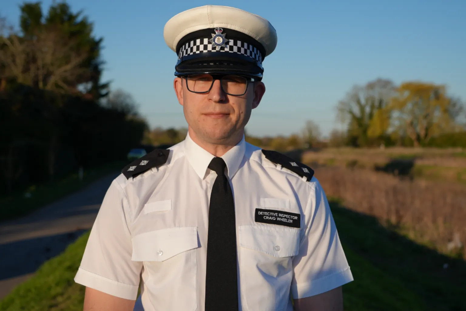 Qays Najm/BBC Det Insp Craig Wheeler is wearing a black and white police cap, a white short-sleeved shirt and a black tie. Behind him is a stretch of road and river. He is looking at the camera.