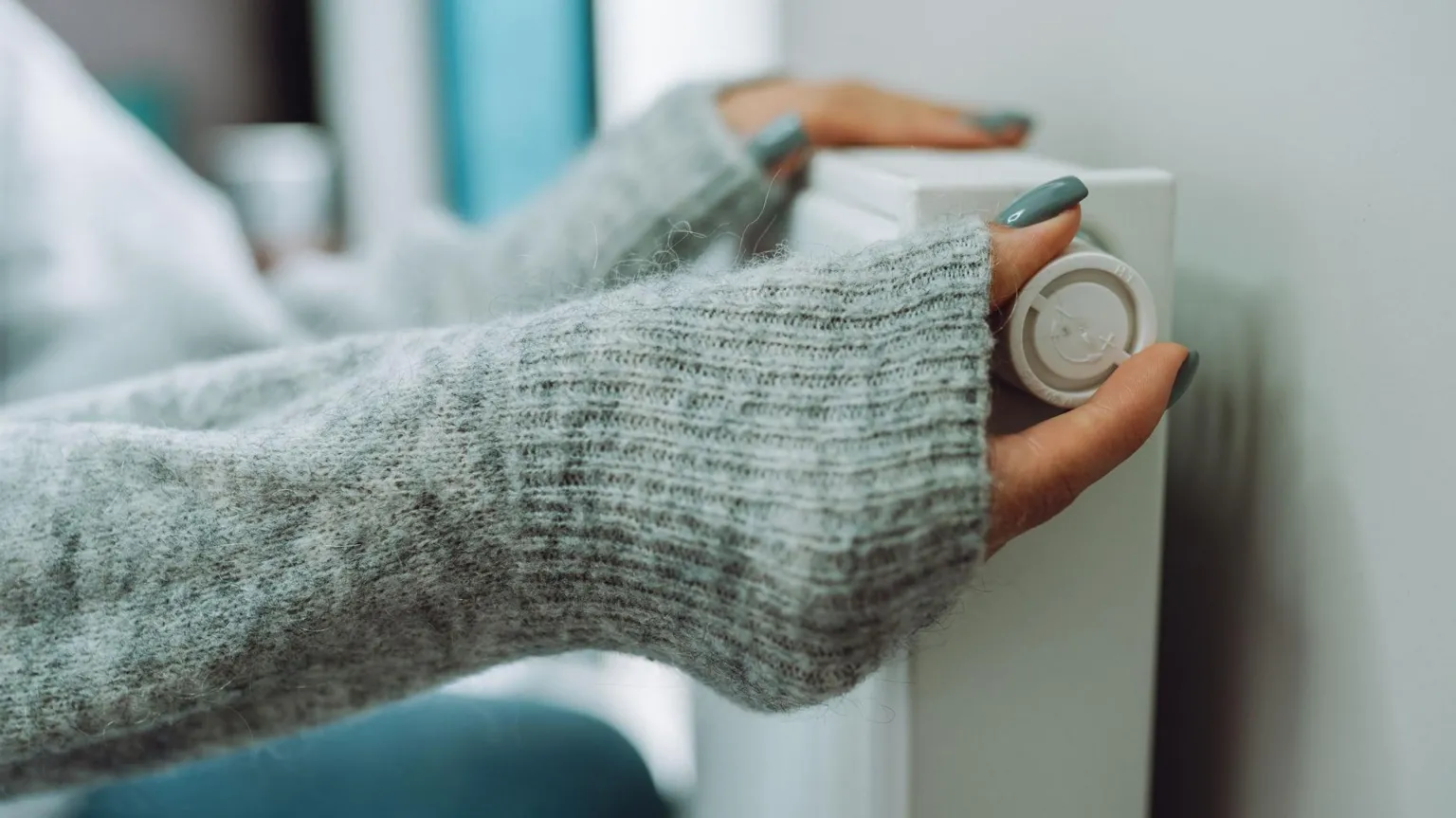  A woman places her hands, which are semi-covered by her grey cardigan, turns a dial on a radiator. The image is a close-up of the womans hands.