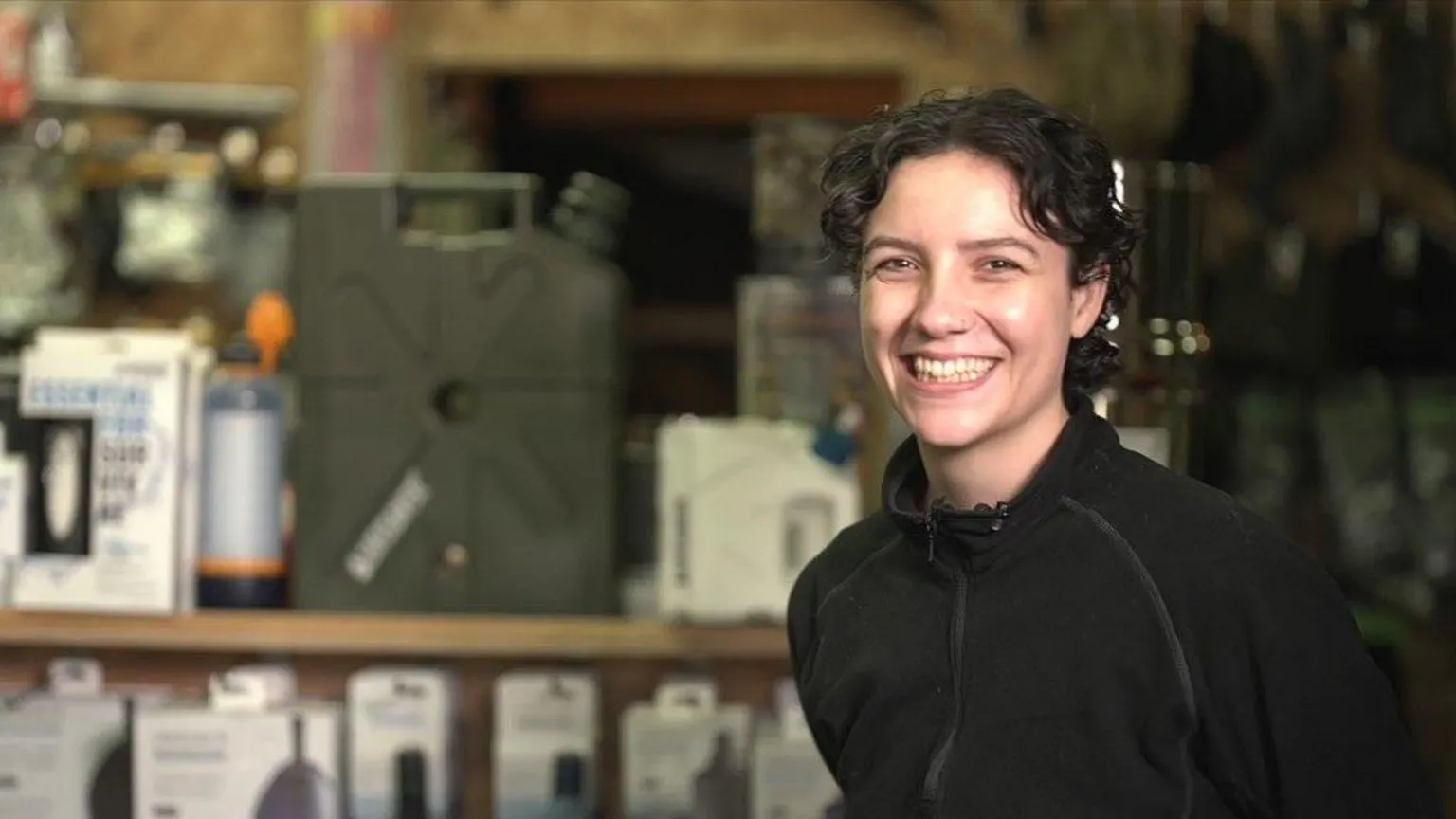 A woman with short brown curly hair grins at the camera. She is stood in a shop full of survival gear. She is wearing a black fleece. 