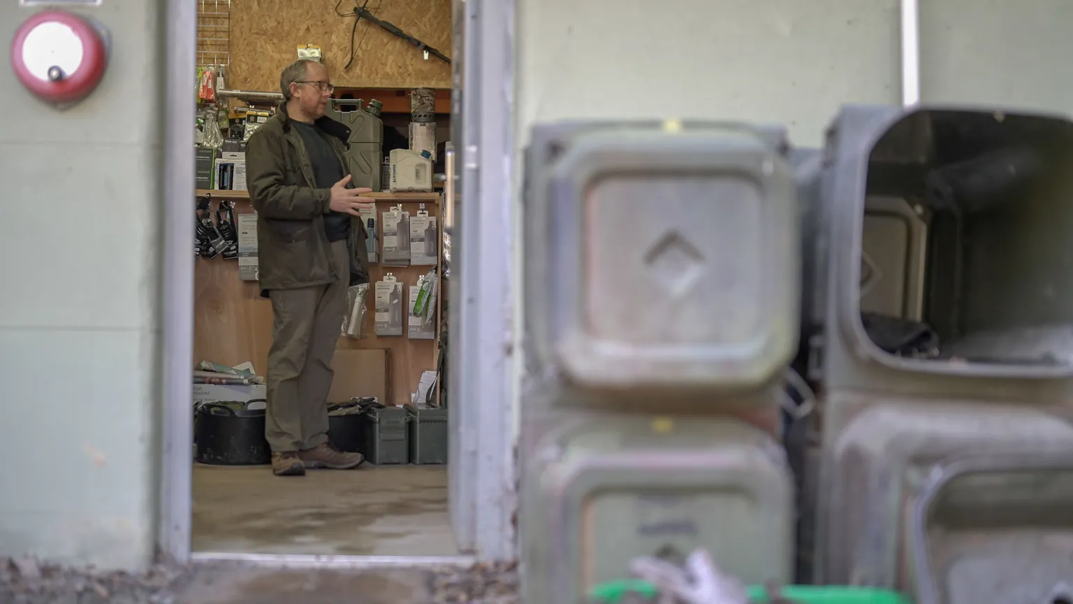 Looking through an open doorway into a workshop‑style shop. Inside, shelves are filled with survival gear, water filters and equipment hanging on the walls. A person stands near the centre of the room, wearing outdoor clothing and gesturing with both hands. In the foreground, outside the doorway, several stacked plastic containers with worn surfaces sit against the wall.