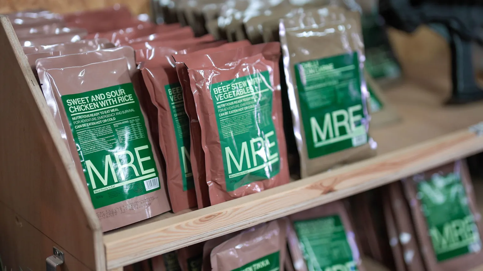 Rows of sealed MRE meal packets sit on wooden shelves. The packets are different colours, including brown and tan, with large green labels. Visible flavours include “Sweet and Sour Chicken with Rice” and “Beef Stew with Vegetables.” The packets are arranged upright in neat lines inside a light‑coloured wooden display unit.