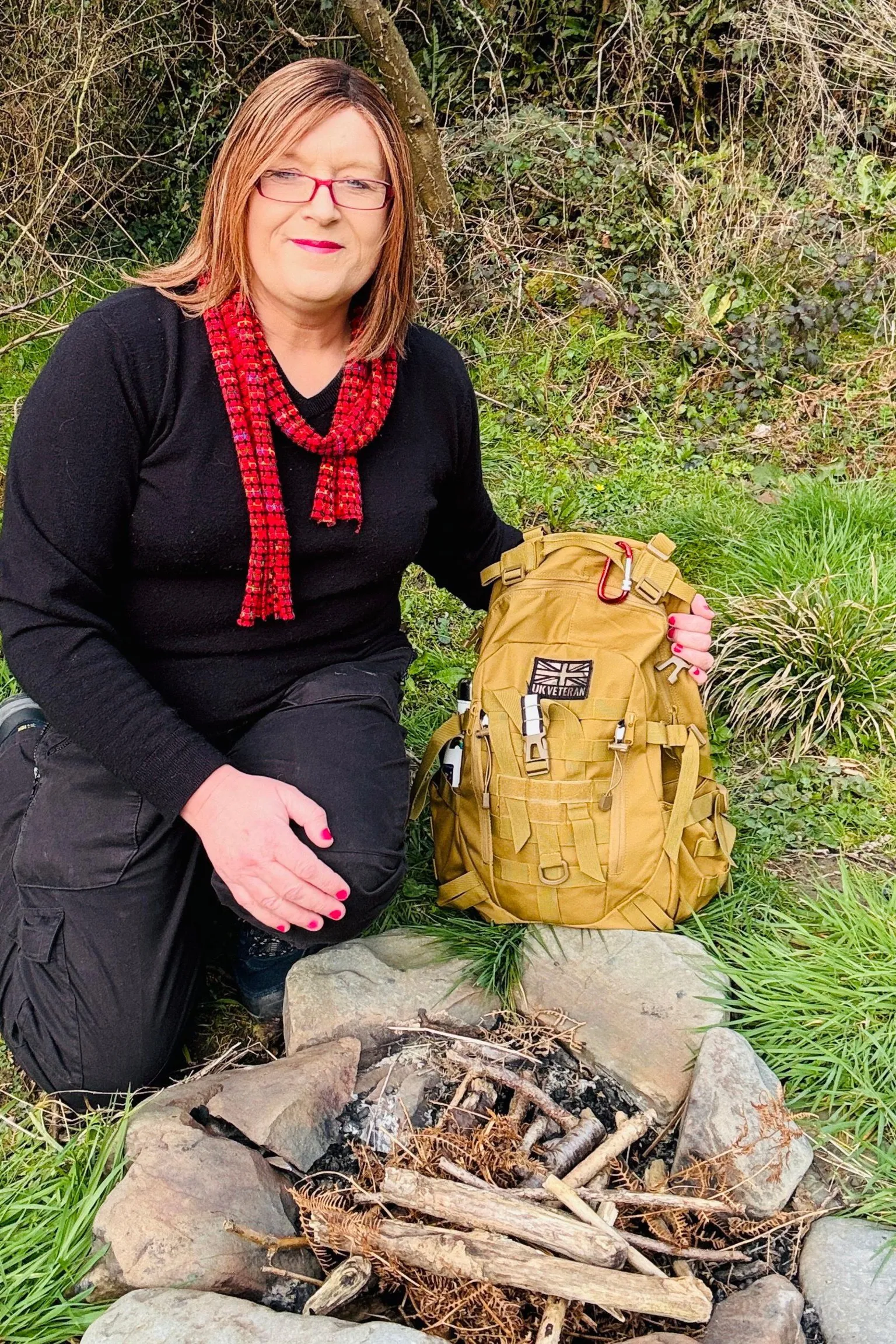 Donna Lloyd A woman with shoulder length brown hair is dressed in black outdoor clothing and is kneeling on grass next to a simple stone firepit containing sticks and charred wood. Beside her is a large tan-coloured rucksack with multiple straps and pockets, resting upright on the ground. Behind the woman is an area of long grass and shrubs, suggesting a woodland or countryside setting.