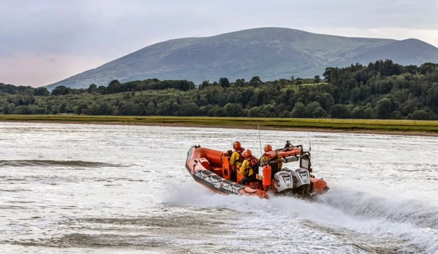 Kim Ayres A lifeboat skipping across the water surface on the Solway estuary with trees and hills in the background