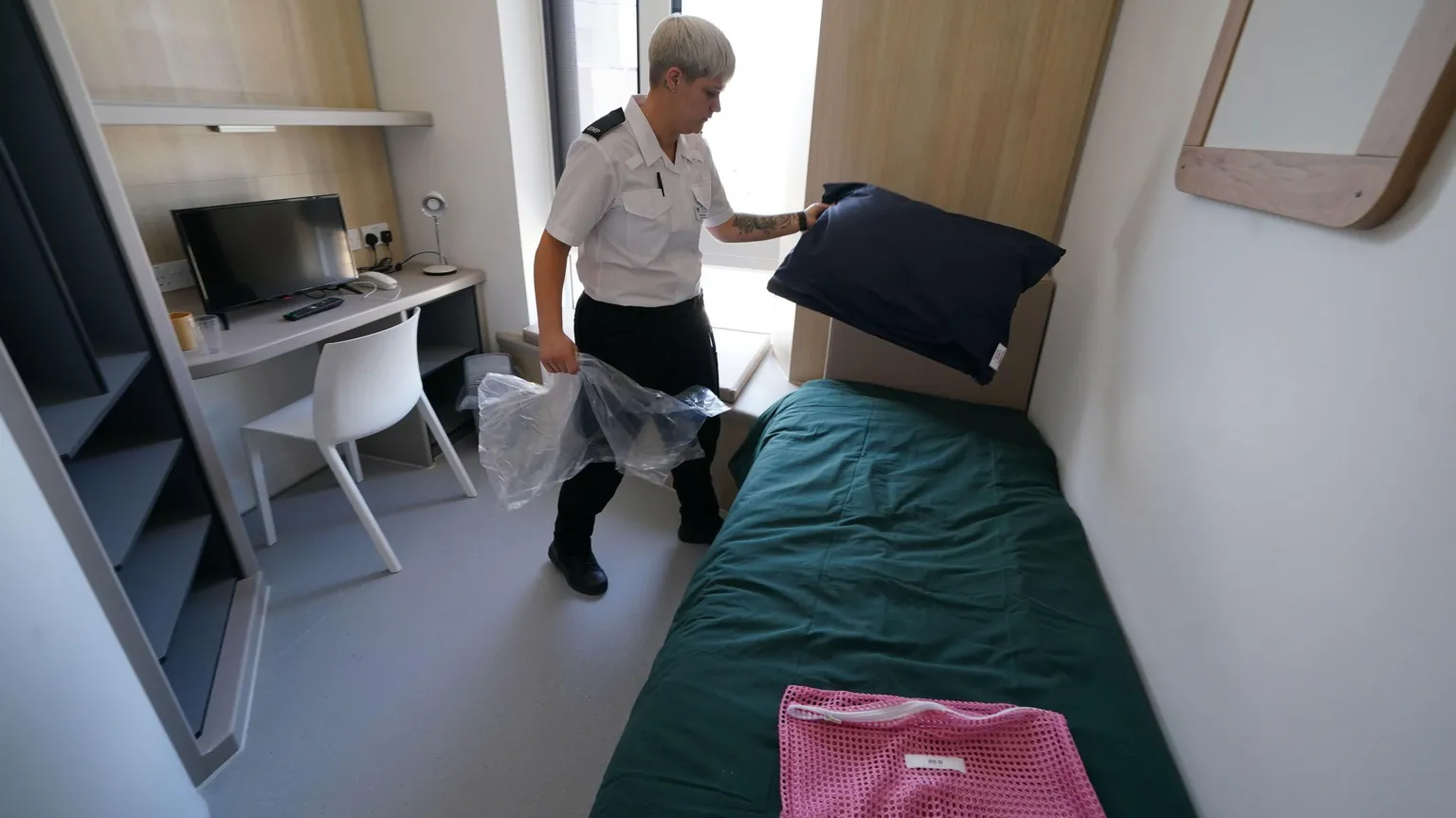  A prison officer adjusts the bedding inside a room within Iris House. The room is small, with a large window, a single bed with dark green bedding opposite a desk and chair with a TV on it.