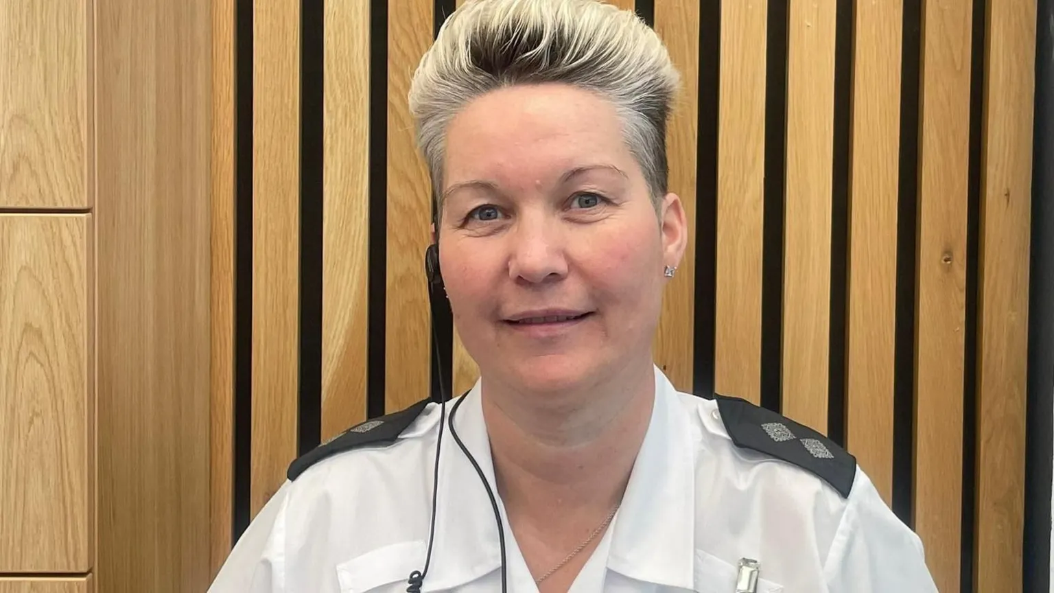 Christina Hogg smiles against the slatted wooden wall behind her. She wears a prison officer's uniform - a white shirt with navy blue epaulettes, a radio earpiece, and has short blonde hair.