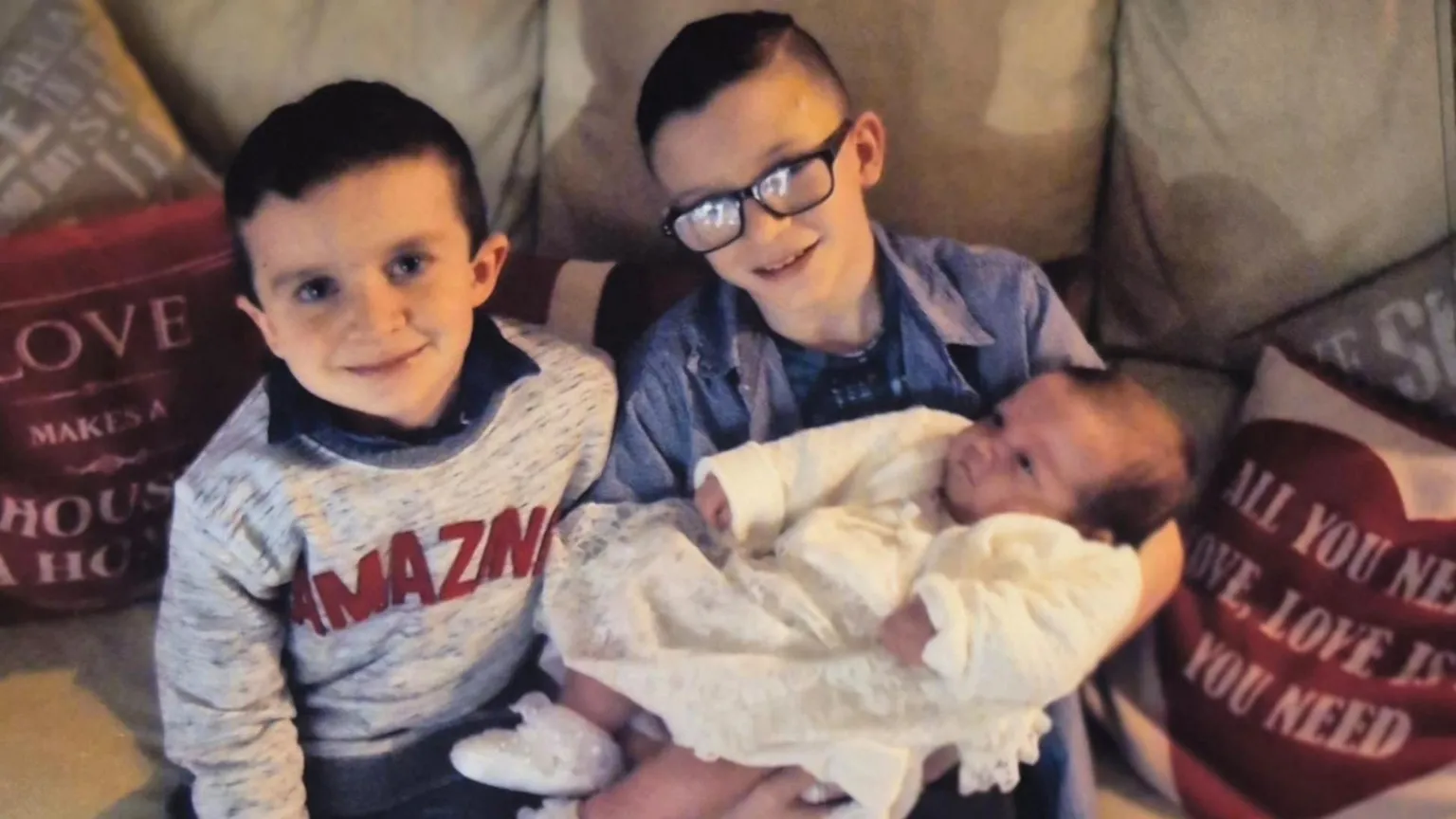 Louise James Evan and Mark with their sister Rioghnach . Evan is wearing a grey sweater with large red letters spelling out AMAZING. He has short black hair. Next to him Mark has black square-rimmed glasses and is wearing a blue shirt. Both boys are sitting on a tan sofa with pink cushions. Mark is holding Rioghnach who is wearing a white dress and cardigan.