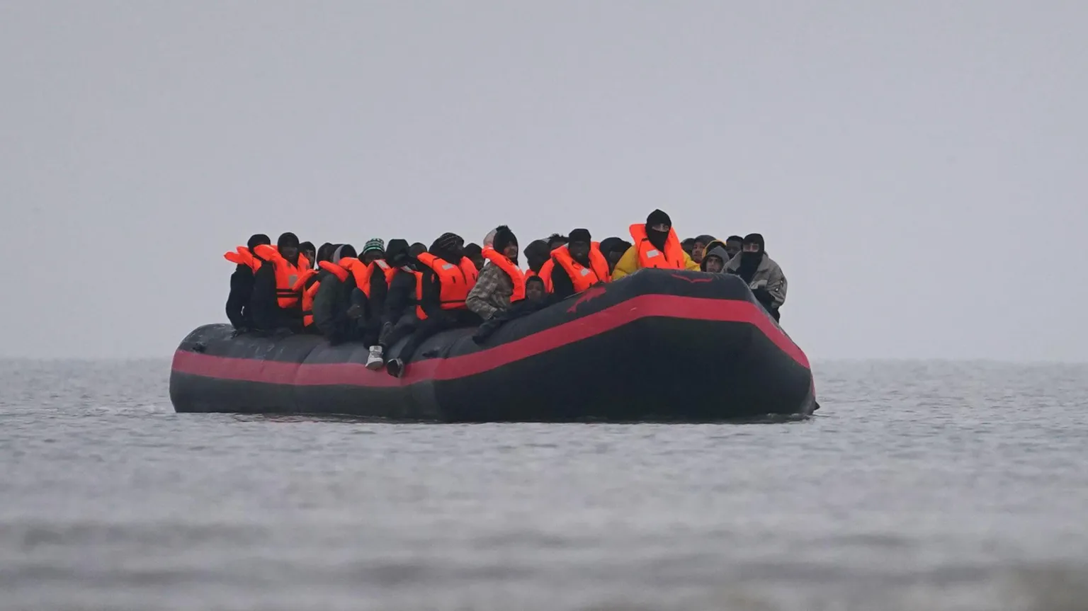  A black dinghy with a red stripe around it, on a grey sea and grey sky. There are lots of people, mainly men, on the dinghy wearing fluorescent life jackets.