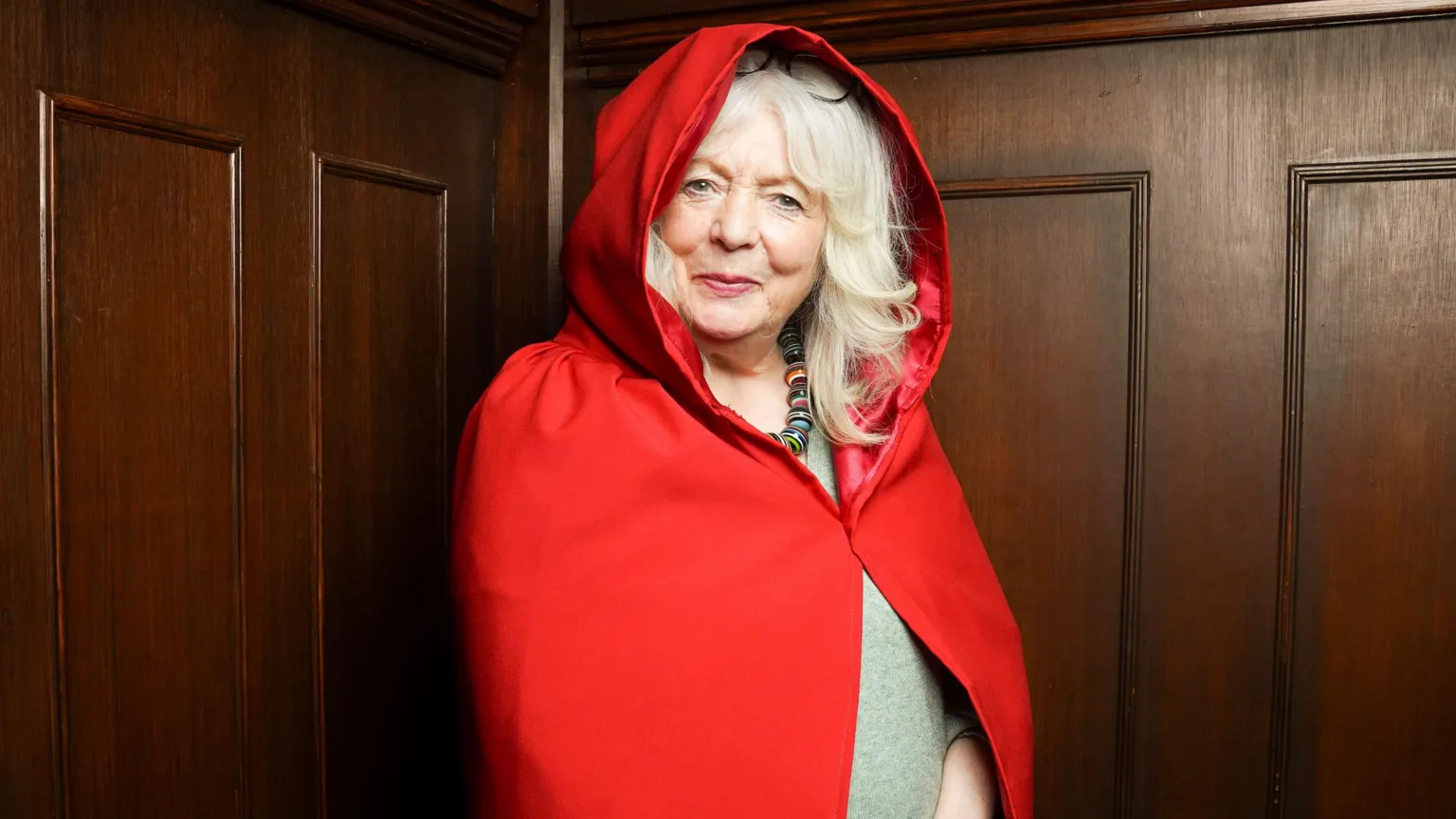Alison Steadman as secret traitor Fiona, she is smiling at the camera conspiratorially and wearing a red hooded cloak.