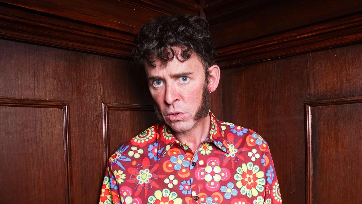 Scott Mills as James, with huge sideburns and a flowery multi-coloured shirt