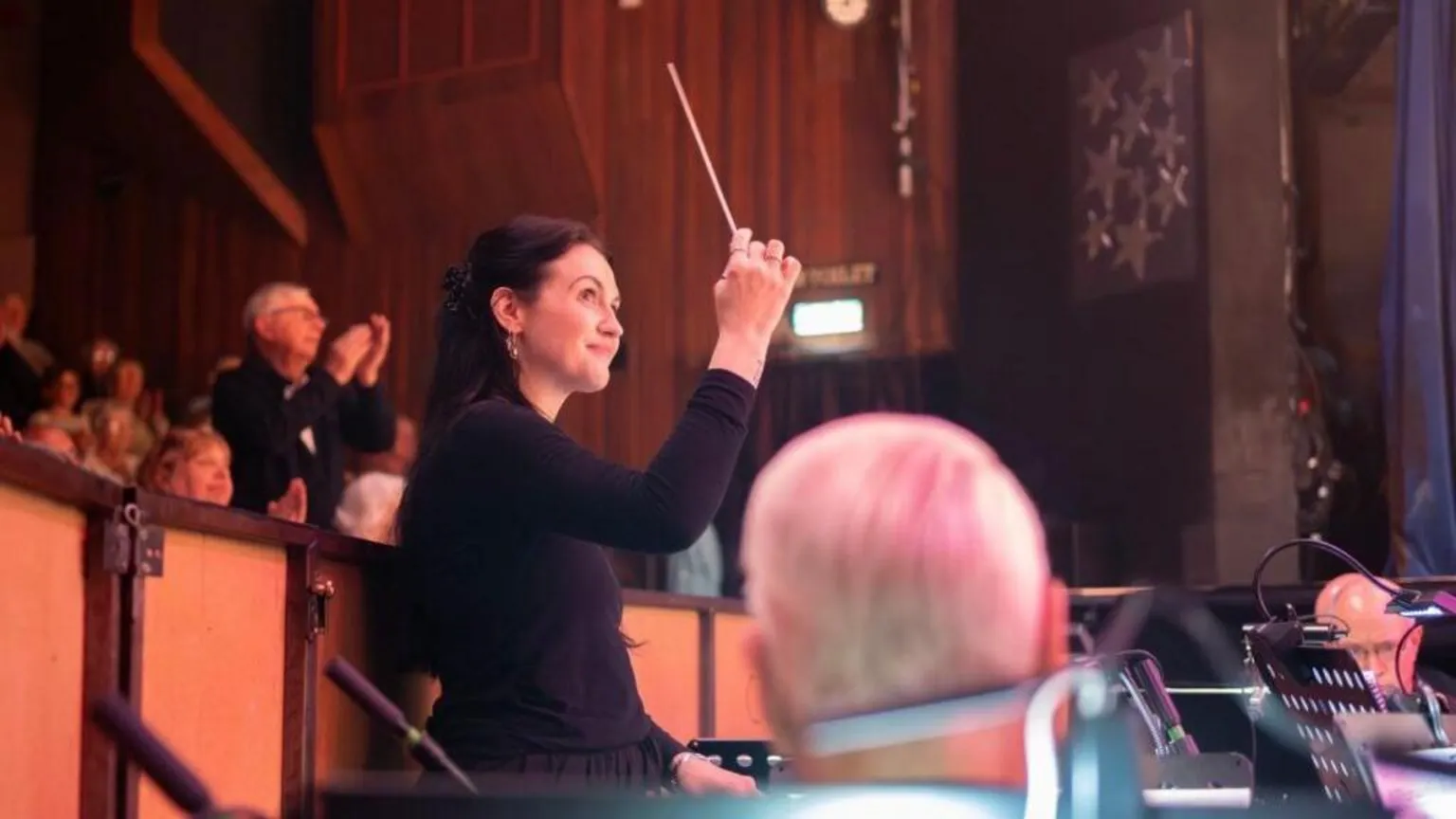 Worcestershire Acute Hospitals NHS Trust Side profile of a woman in black conducting an orchestra. She is holding a baton and smiling. There are people clapping behind her.