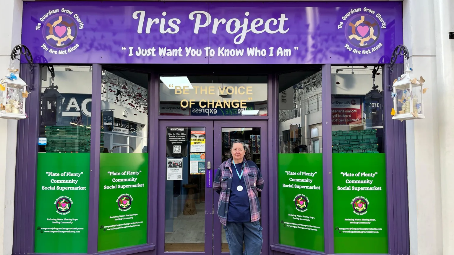 Sioux Watkins is stood in front of the Plate of Plenty shop. She has grey hair tied in a pony tail with glasses on her head. She is wearing a tartan shirt, with a navy T-shirt underneath and blue cargo trousers. 