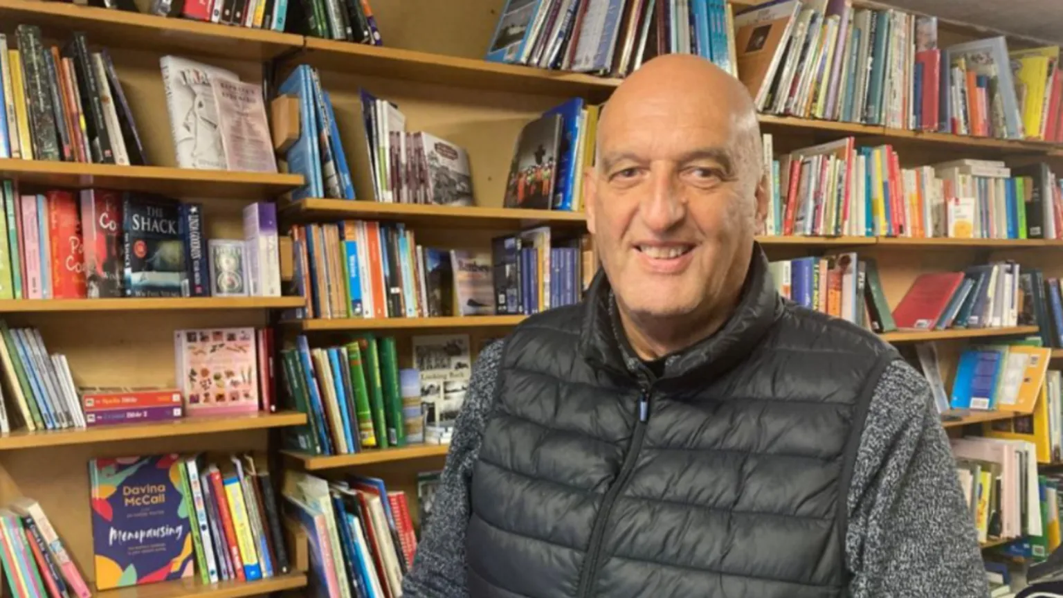 A bald middle aged man wearing a grey jumper and black gillet jacket. He is stood in front of a packed bookshelf and is smiling as he looks at the camera.