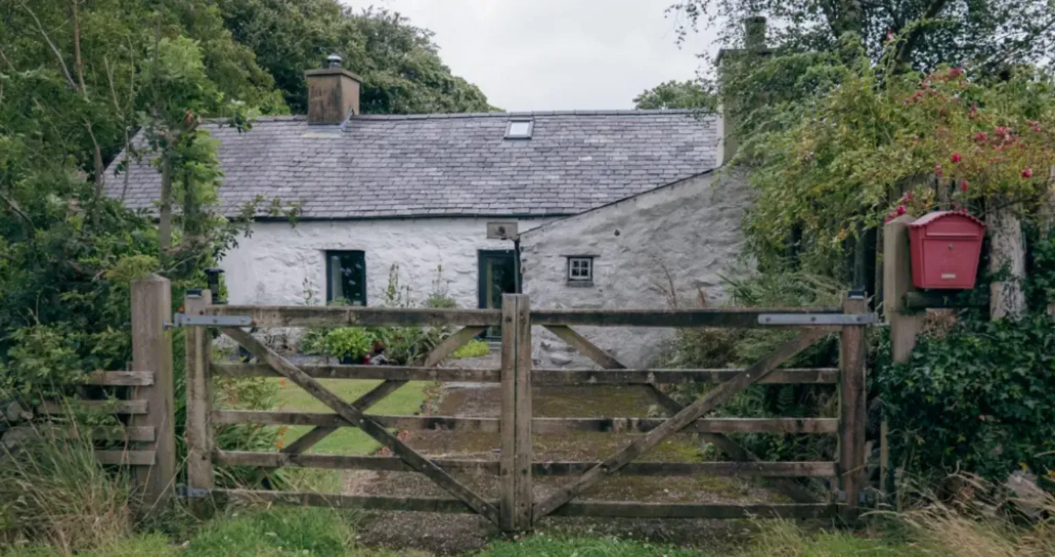 The outside of a white-washed cottage, with grass in front and a wooden gate, with a red post box beside it.