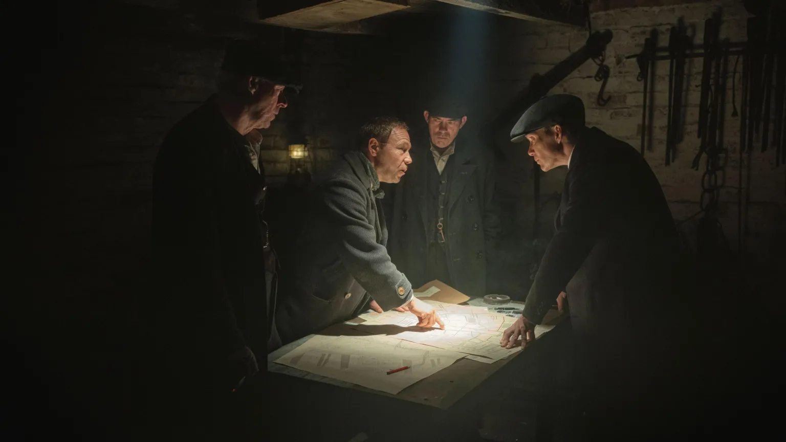 Netflix Several men in old fashioned suits are standing around a table in a basement and appear to be looking at a map which is lit up by an overhead light. One of the men on the left is pointing at the map.