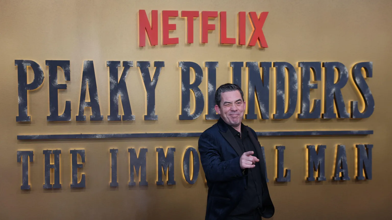 EPA Packy Lee in a dark suit with black hair is winking and pointing toward the camera. He is standing in front of a a large gold sign which says: 'Netflix Peaky Blinders The Immortal Man'